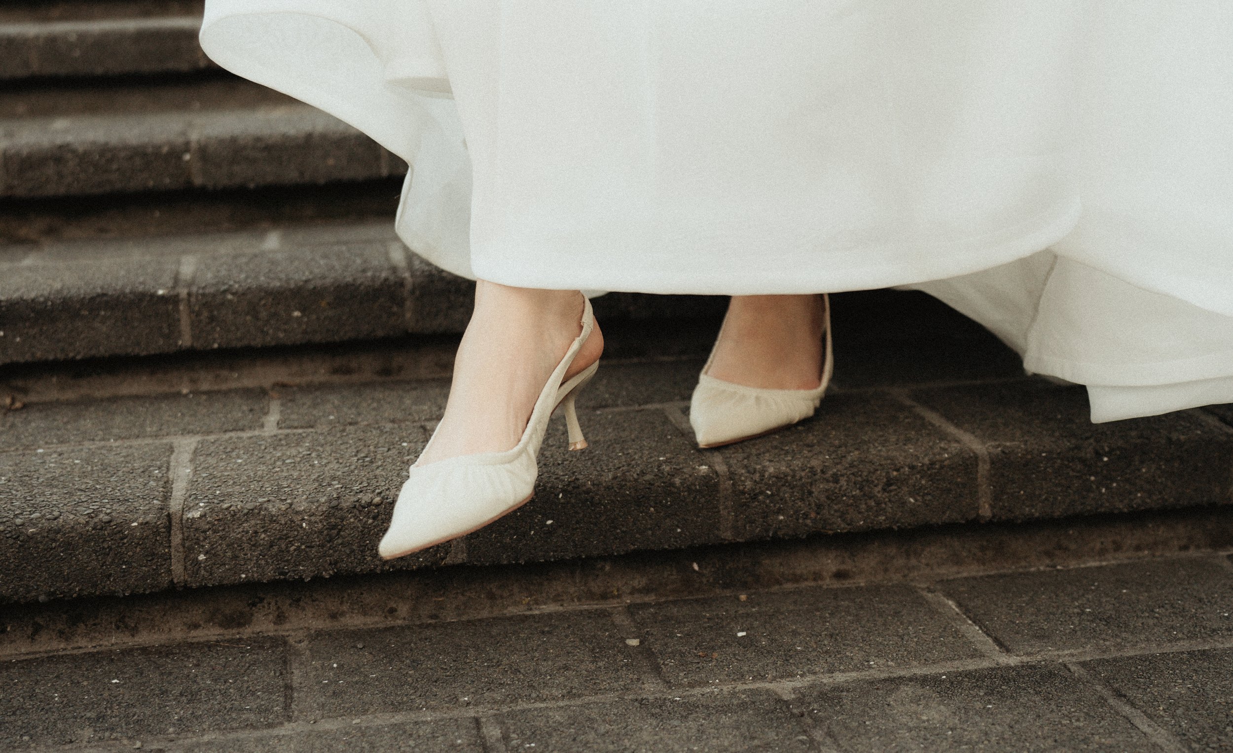 Close-up of a woman's feet wearing white high-heeled shoes beneath a white gown, standing on a brick step.