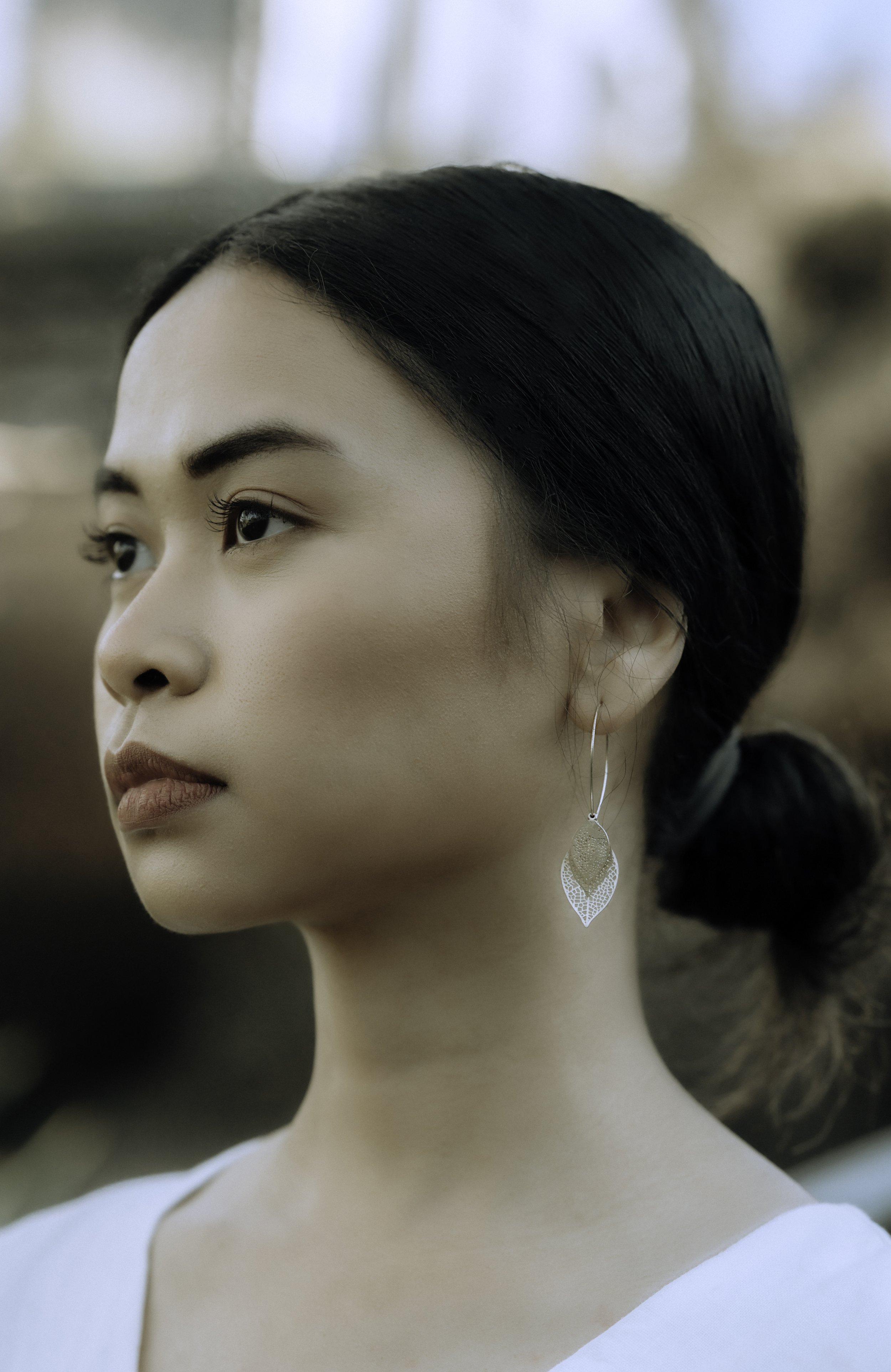 Close-up of a woman with dark hair pulled back, wearing a white top and floral earring, with a blurred background.