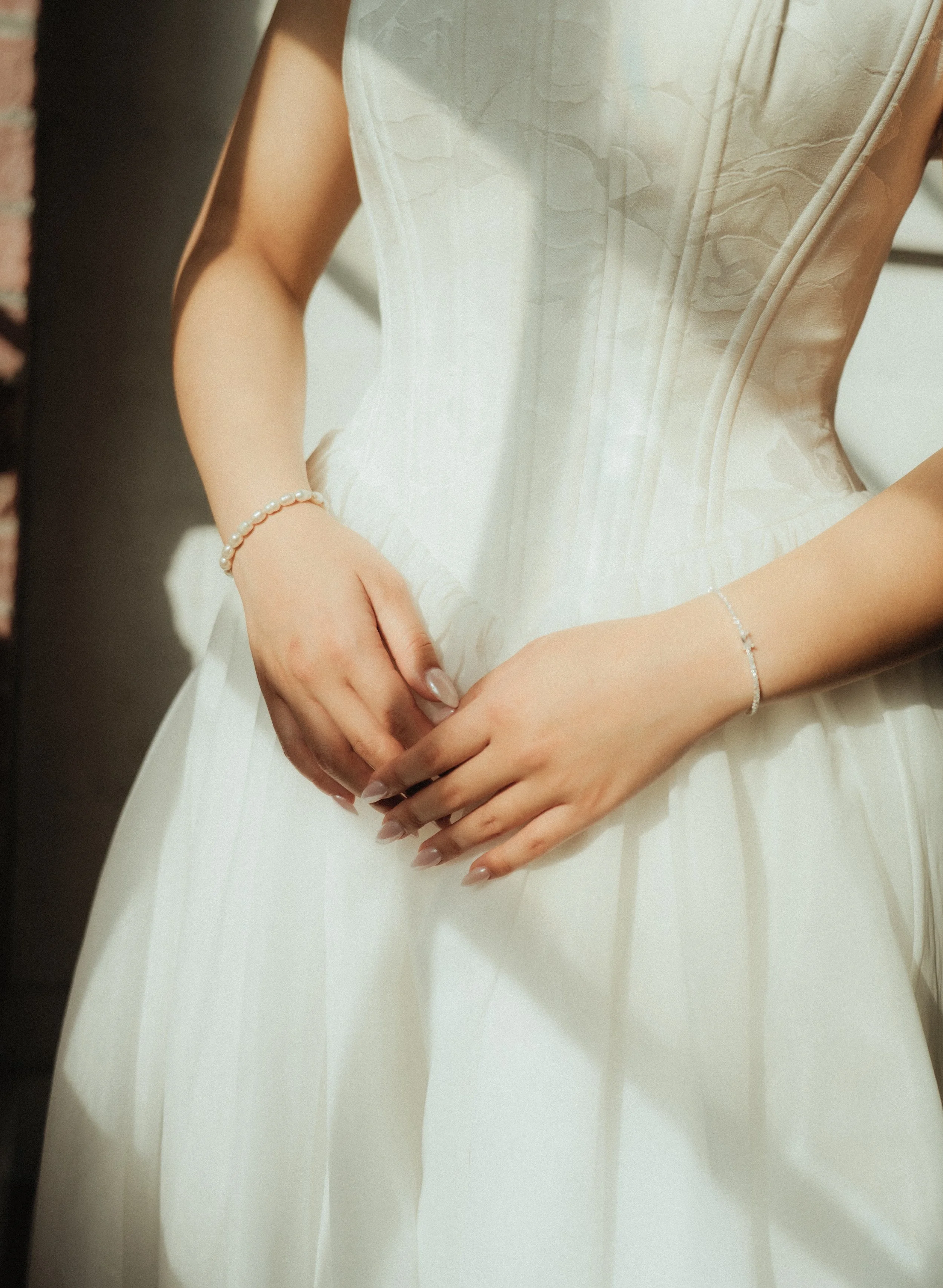 Close-up of a bride's hands resting on her lap, wearing pearl and diamond jewelry, in a white wedding dress with delicate embroidery.