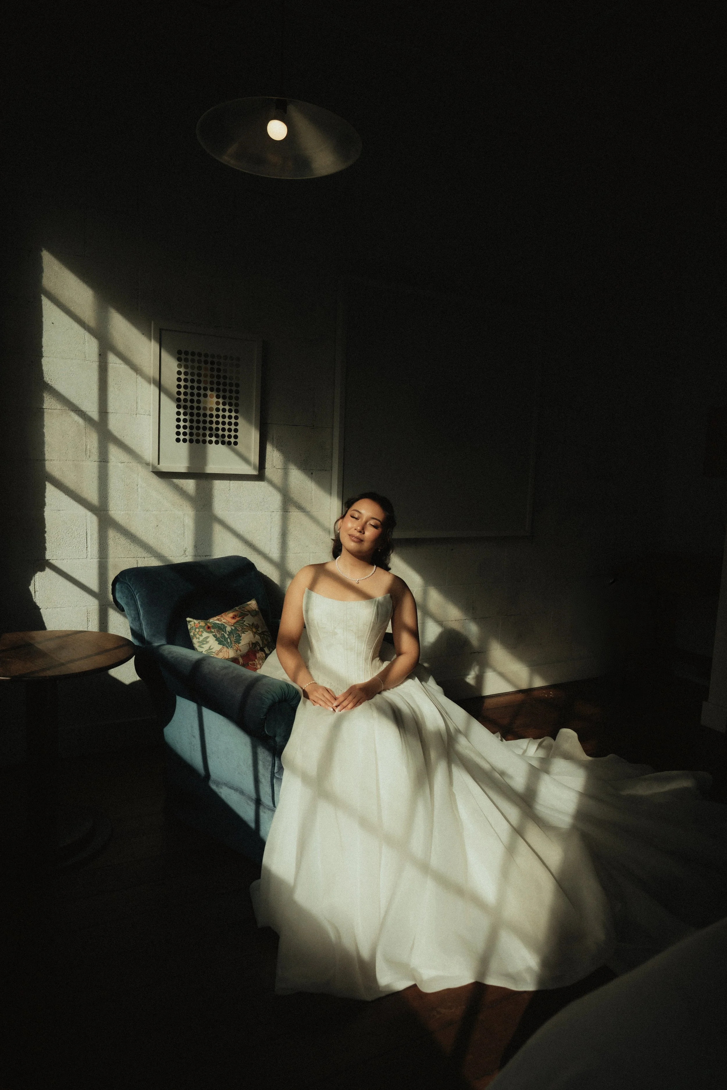 A woman in a wedding dress sitting on a blue couch in a dimly lit room with sunlight casting shadows through a window.