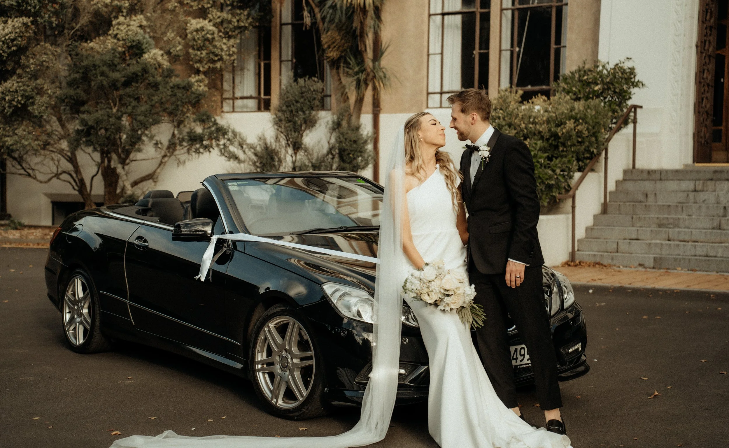 A bride and groom standing close together in front of a black convertible car, smiling and touching foreheads. The bride is holding a bouquet of white flowers, wearing a white wedding dress and veil. The groom is in a black tuxedo with a white shirt 