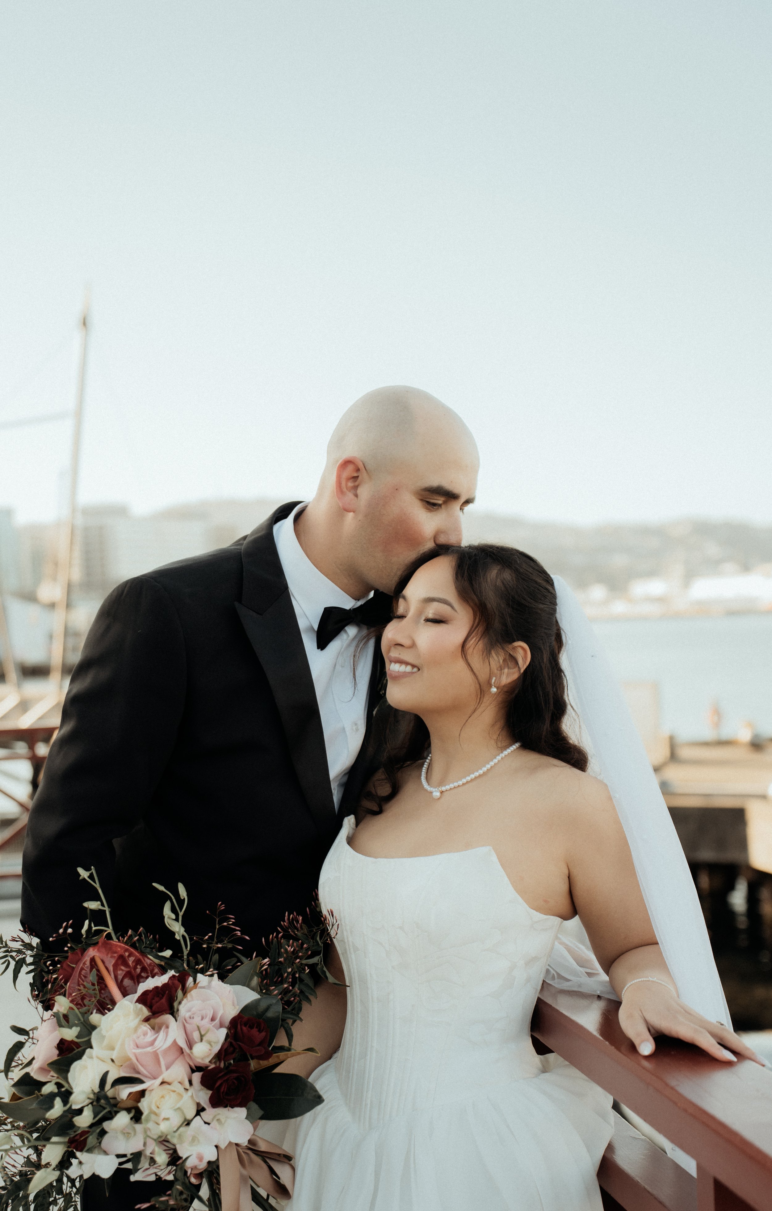 A newlywed couple on a bridge outdoors, the groom in a black tuxedo and the bride in a white strapless wedding dress, holding a bouquet of pink, white, and maroon flowers, with water and city buildings in the background.