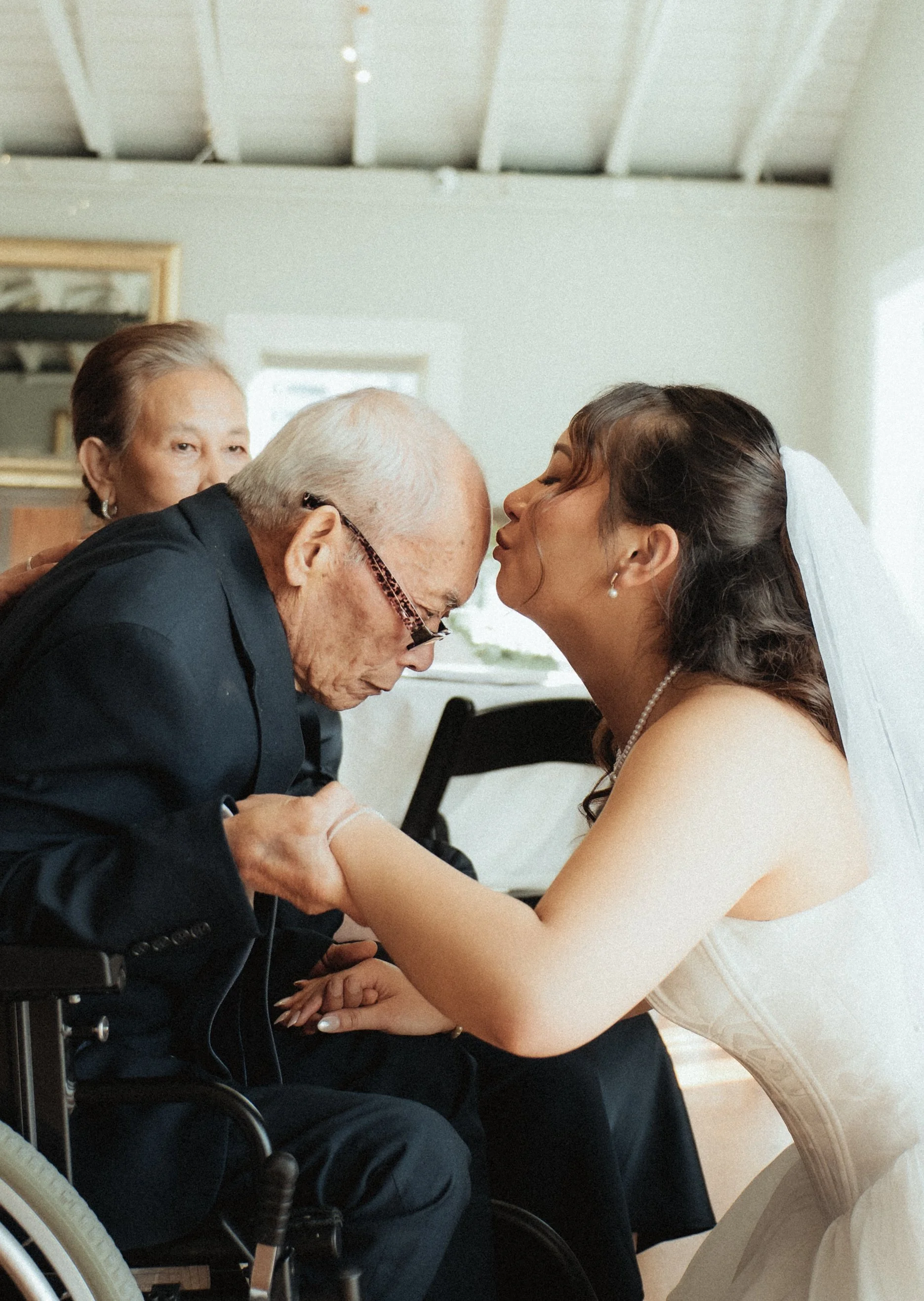 A bride and an elderly man are sharing a kiss at a wedding. A woman is watching in the background.