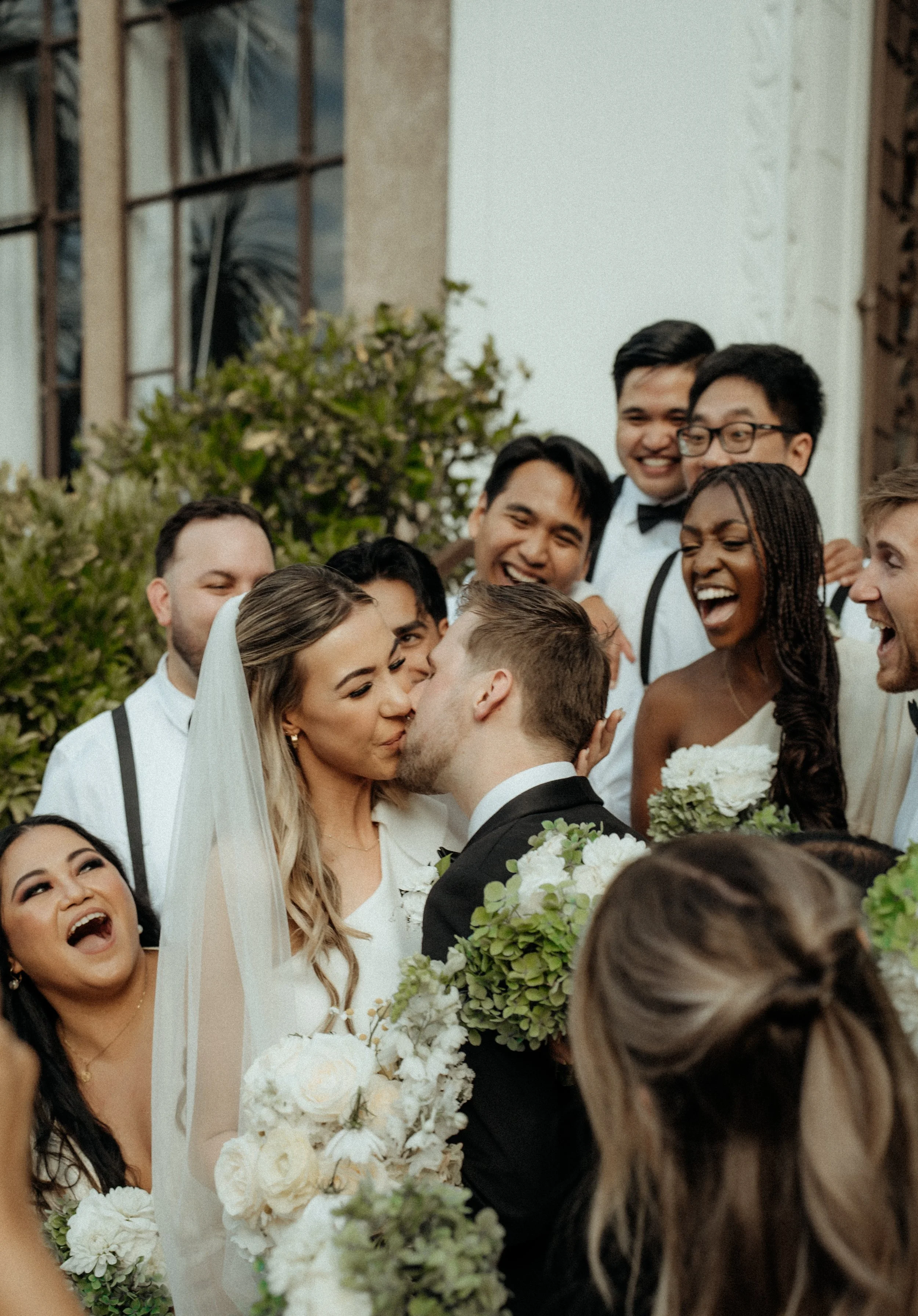 The bridal party gathered outdoors, smiling and celebrating as a bride and groom kiss in the foreground, surrounded by bouquets of white flowers and greenery.
