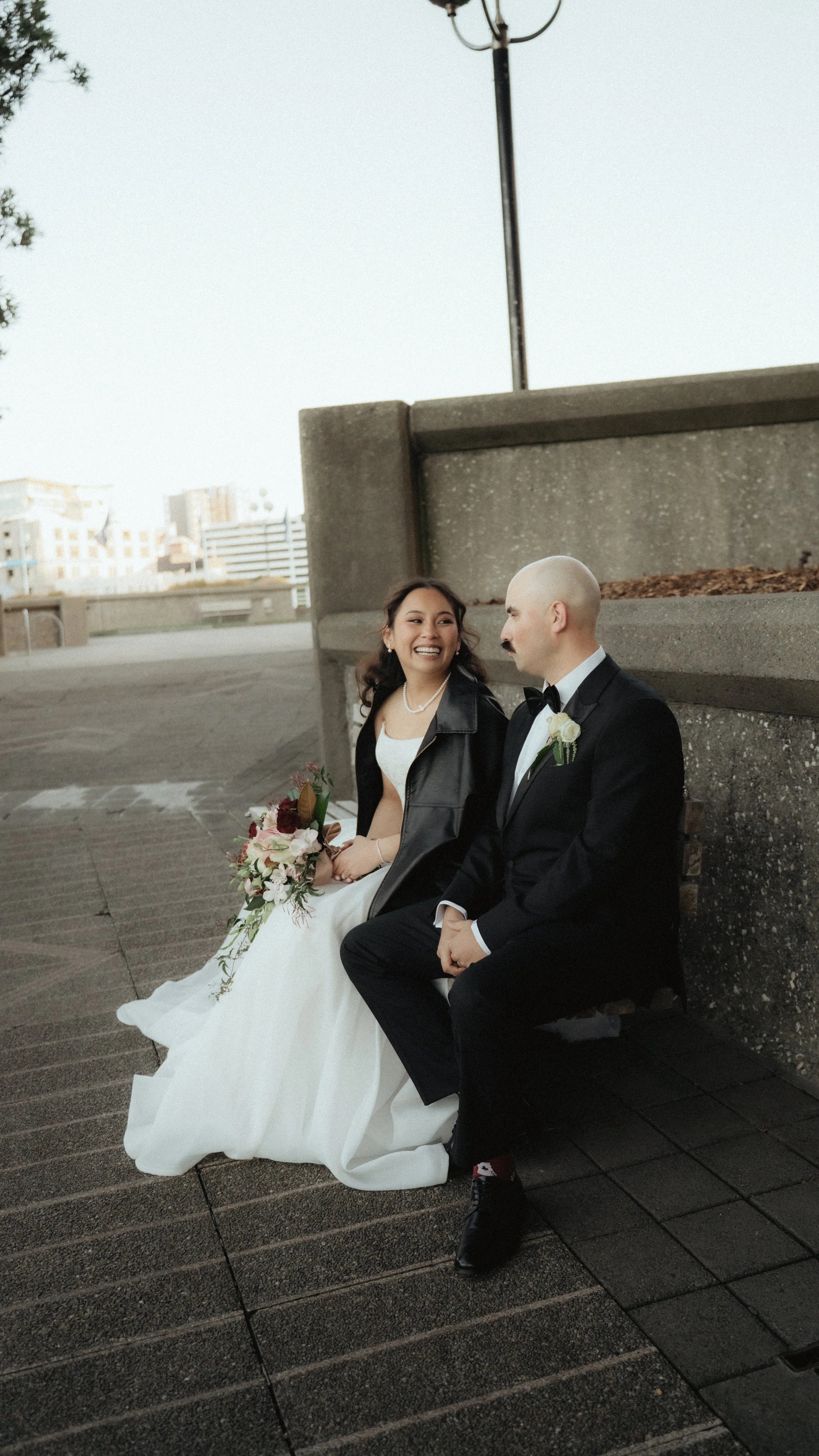 A bride and groom sitting on a bench outdoors, smiling and looking at each other. The bride wears a white wedding gown and holds a bouquet of flowers. The groom is dressed in a black tuxedo with a bow tie, a white rose boutonniere, and has a bald hea