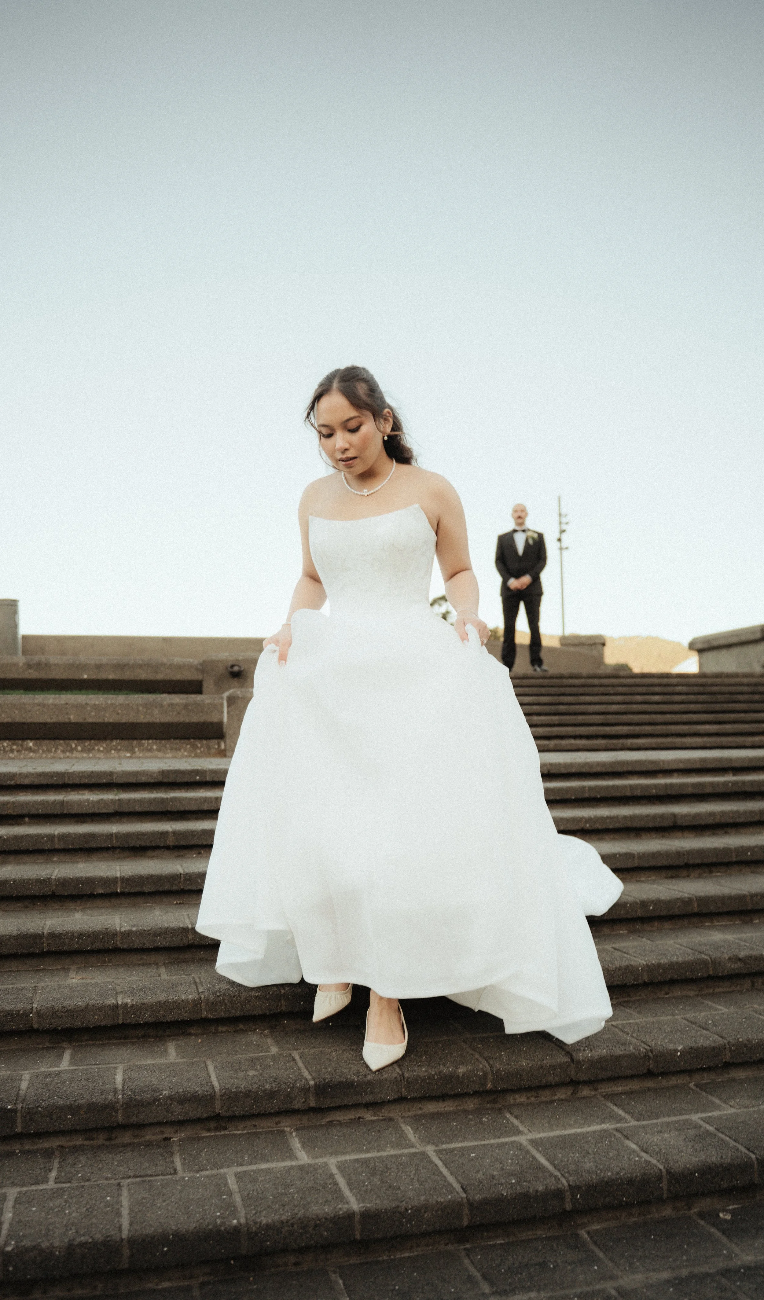 A bride in a white wedding gown standing on outdoor stone steps, with a groom in a black tuxedo and bowtie in the background.