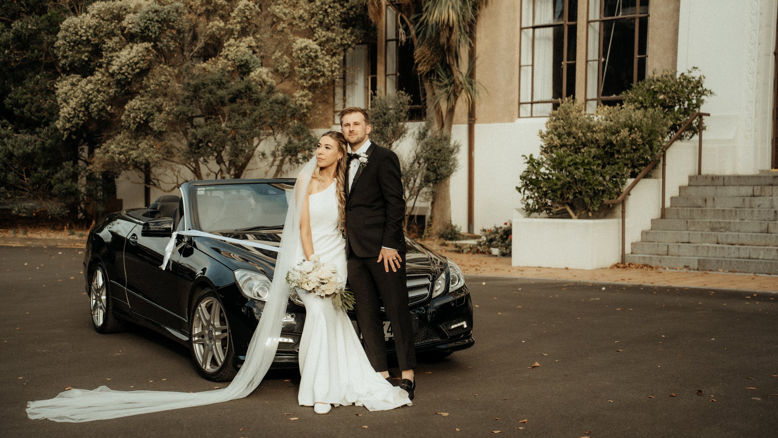 A bride and groom standing beside a black luxury convertible car decorated with ribbons, in front of a building with steps and lush greenery.