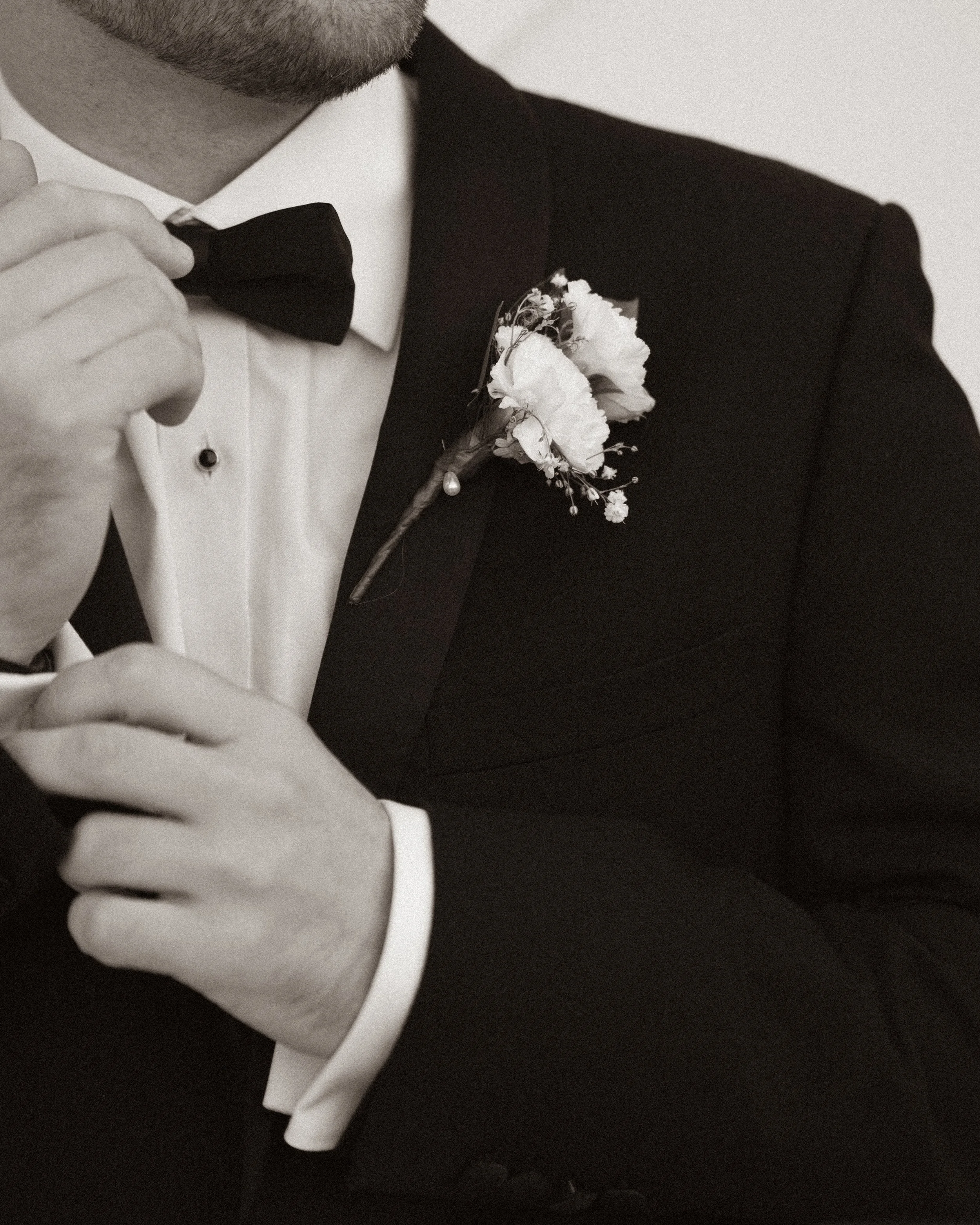 A man in a tuxedo with a boutonnière on his lapel, adjusting his black bow tie with his right hand.