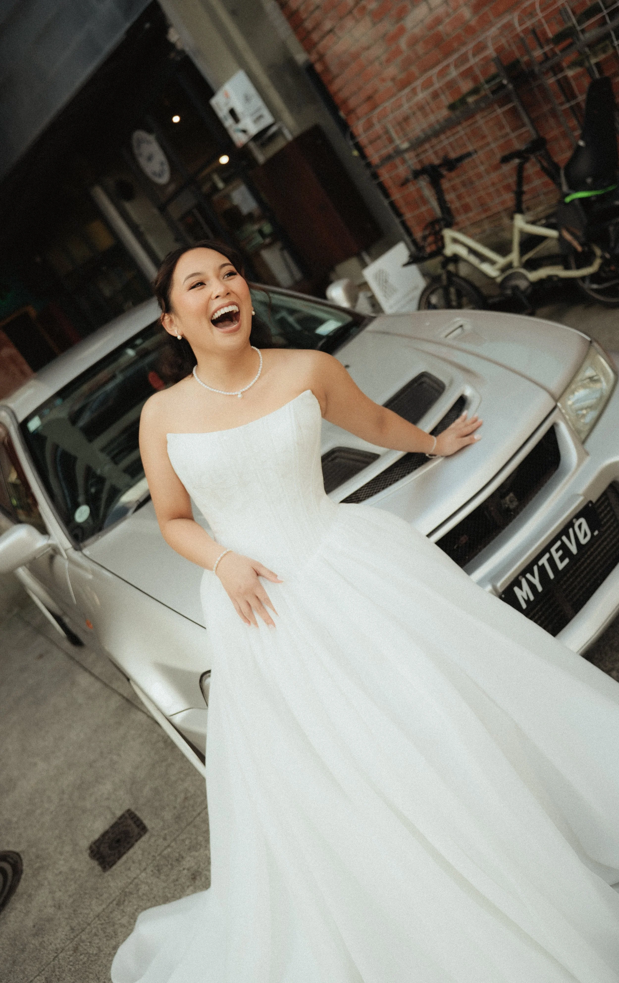 A woman in a white wedding dress standing outdoors, laughing with her hand resting on a white car.