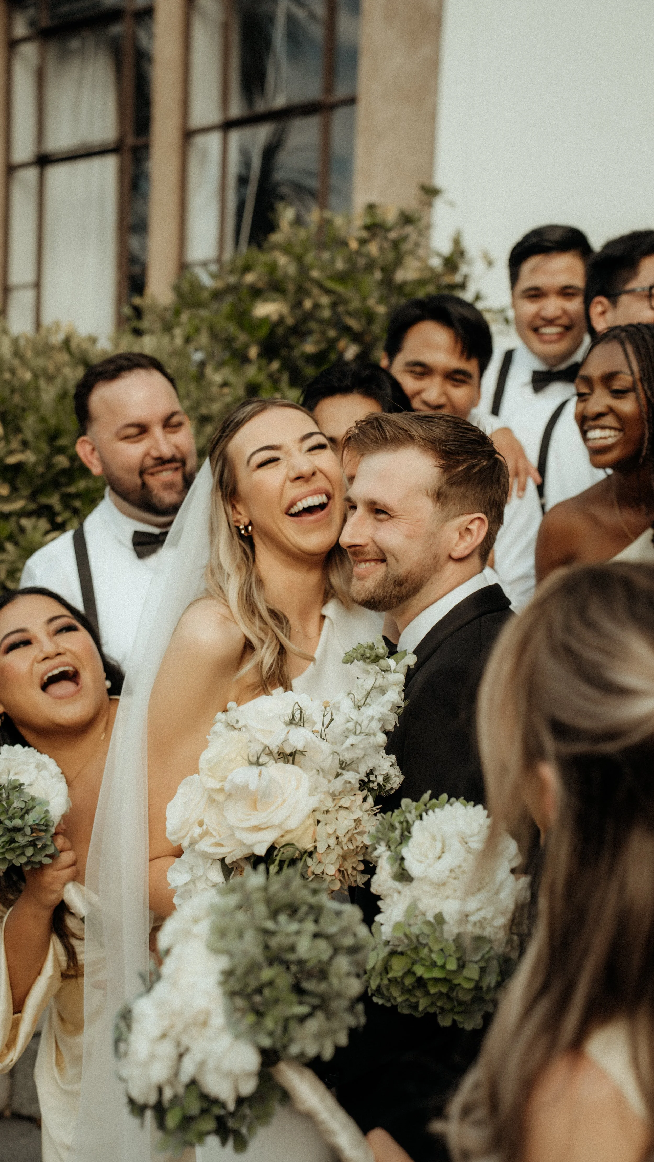 Bridal party, including a bride and groom, smiling and gathered together outdoors for a celebration, with some holding bouquets of flowers.