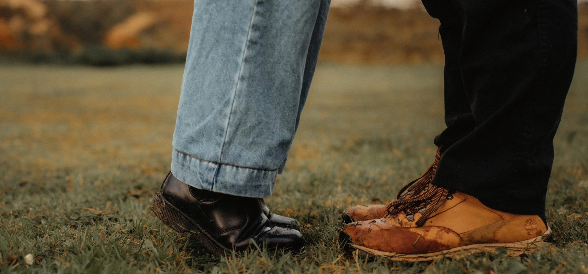 Close-up of two people standing close, showing their shoes and pants on grass; one person in black pants and brown boots, the other in blue jeans and black shoes.