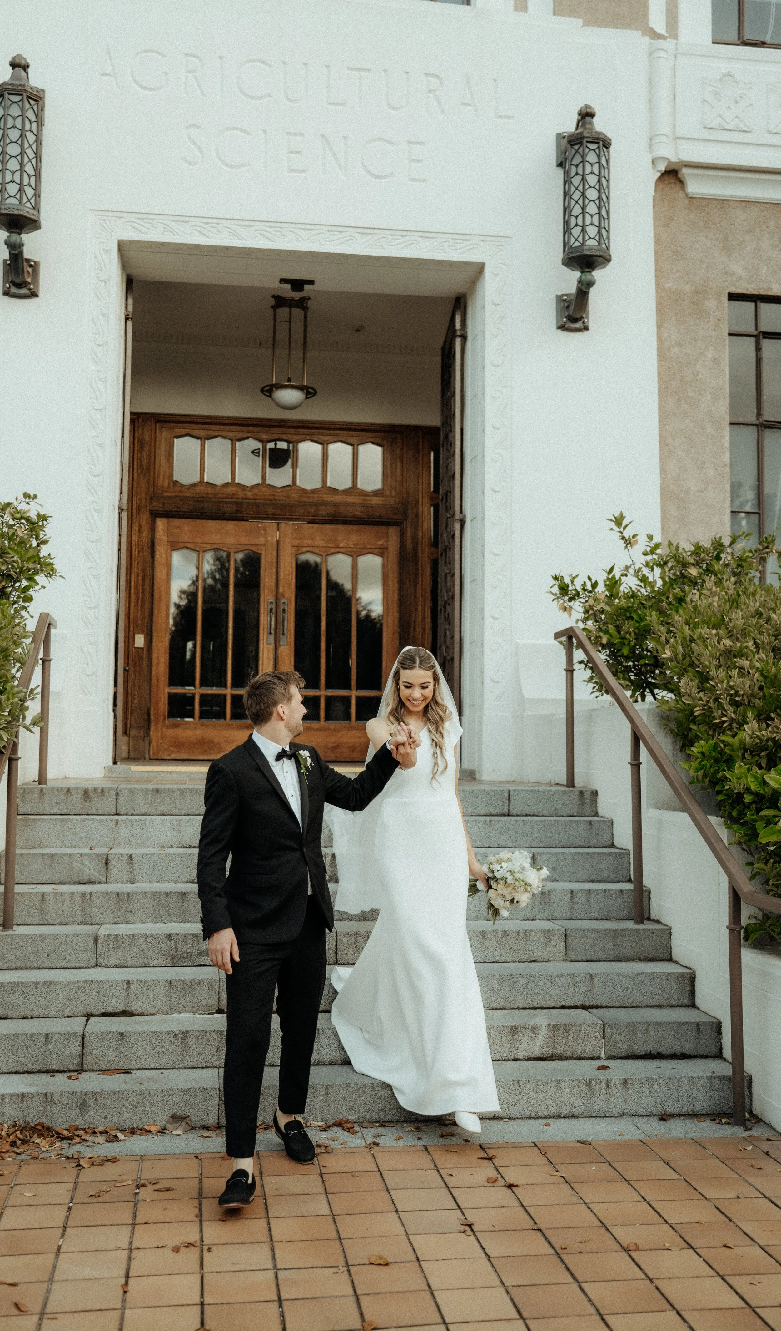 A bride and groom walking down the steps of a building labeled 'Agricultural Science.' The bride is wearing a white wedding dress and holding a bouquet of flowers, while the groom is dressed in a black tuxedo. They are smiling and holding hands, cele