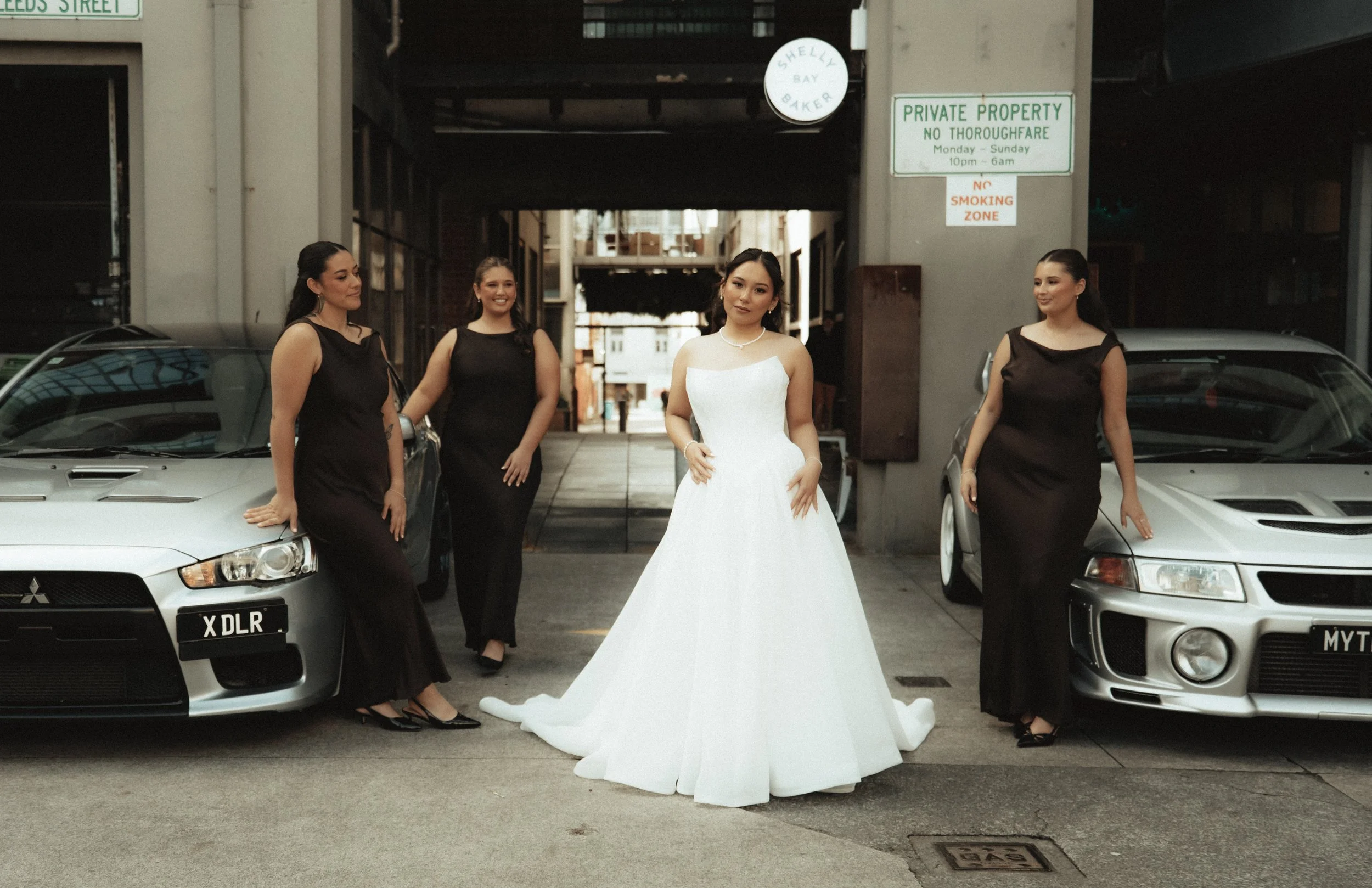 A bride in a white wedding gown stands between four women in black dresses, in a parking lot with parked cars on either side, under an overpass. There are signs about private property and no smoking.