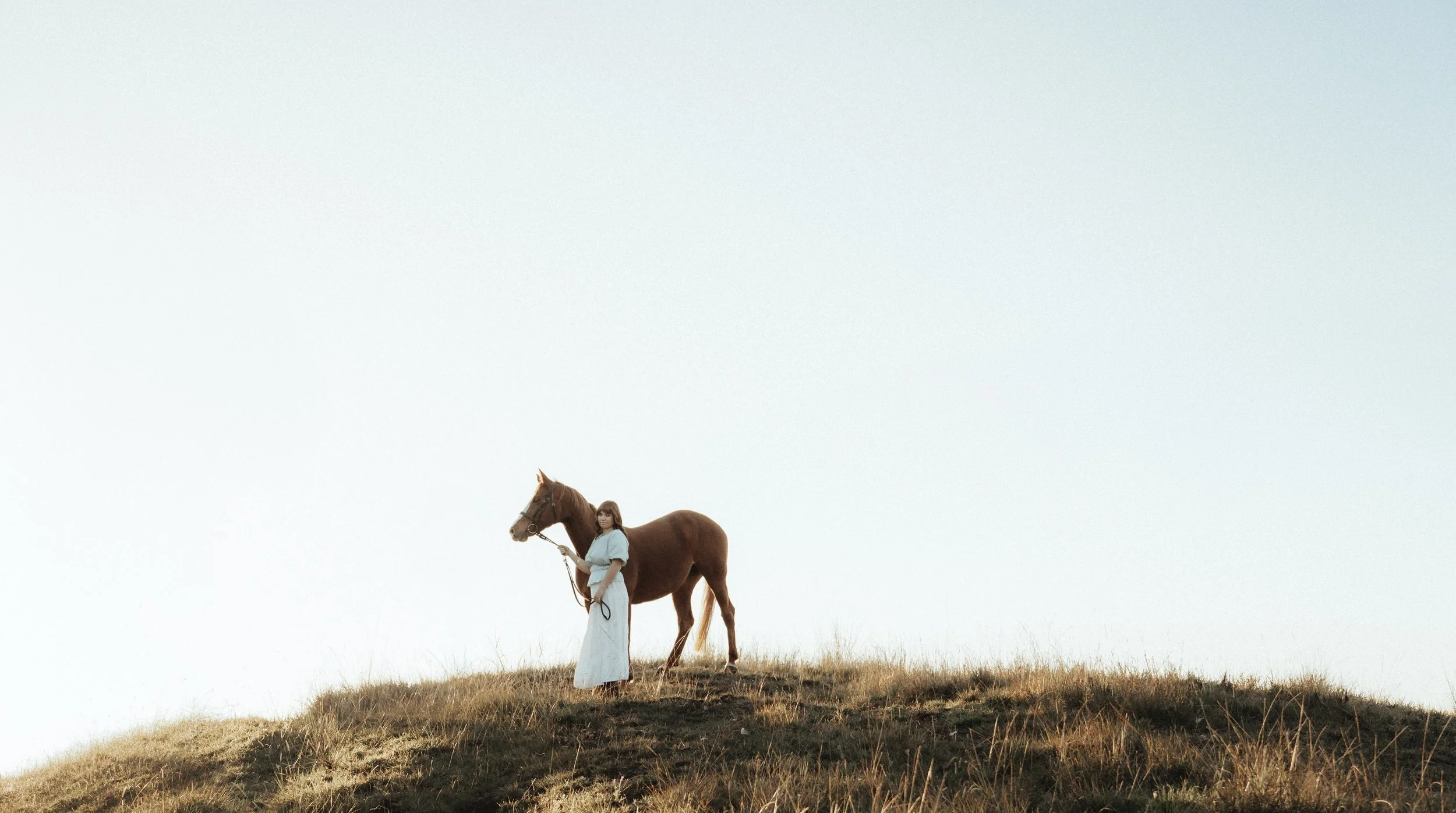 A woman in a white dress holding a lead rope, standing next to a brown horse on a grassy hill under a clear sky.