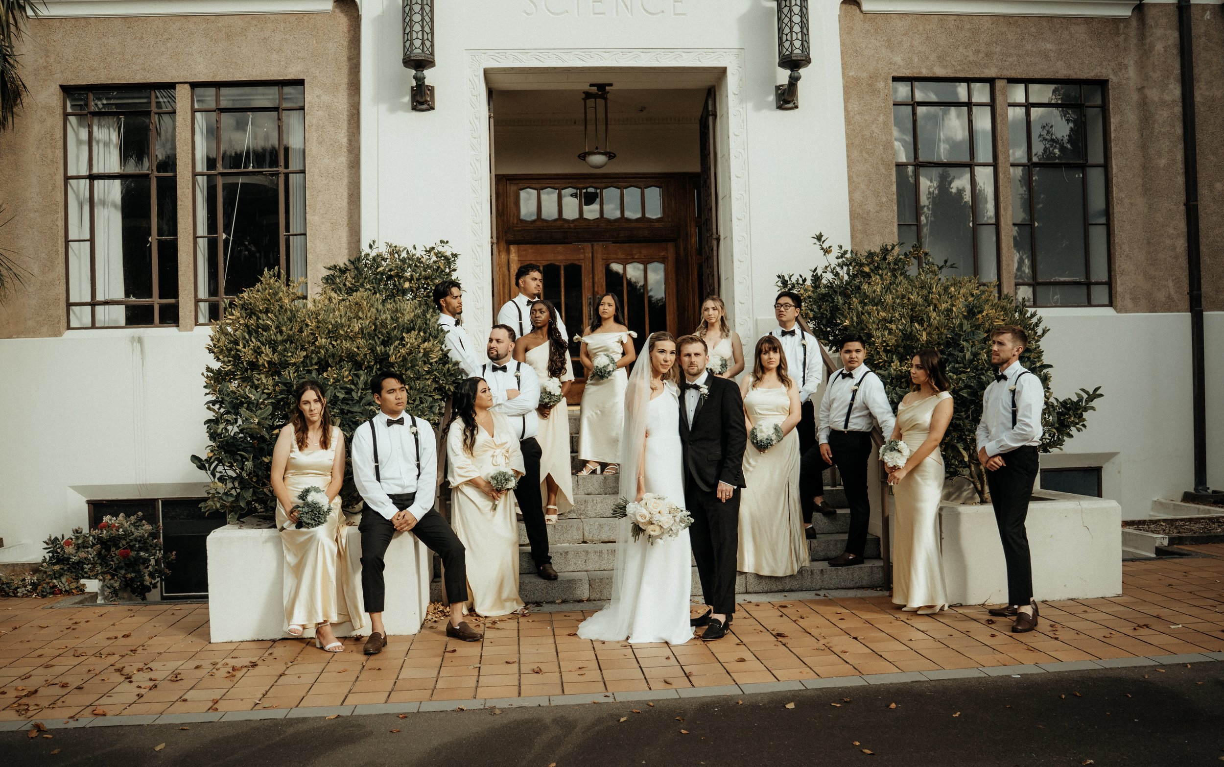 The bridal party, the bride and groom standing on the steps outside a building with large windows and greenery, posing for a wedding photo.