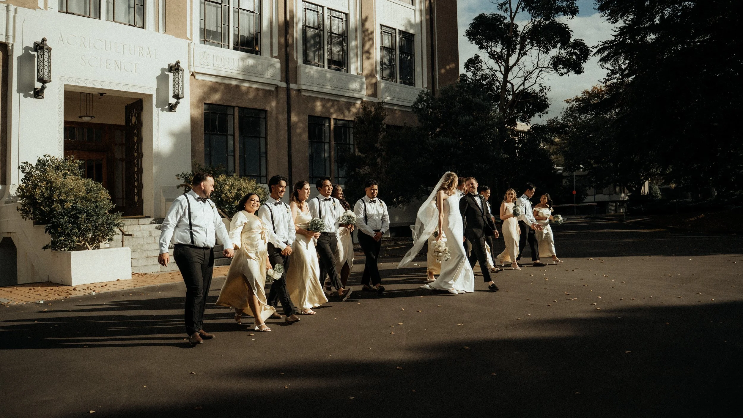 The bridal party dressed in wedding attire walking outside Massey University building called 'Agricultural Science' on a sunny day.