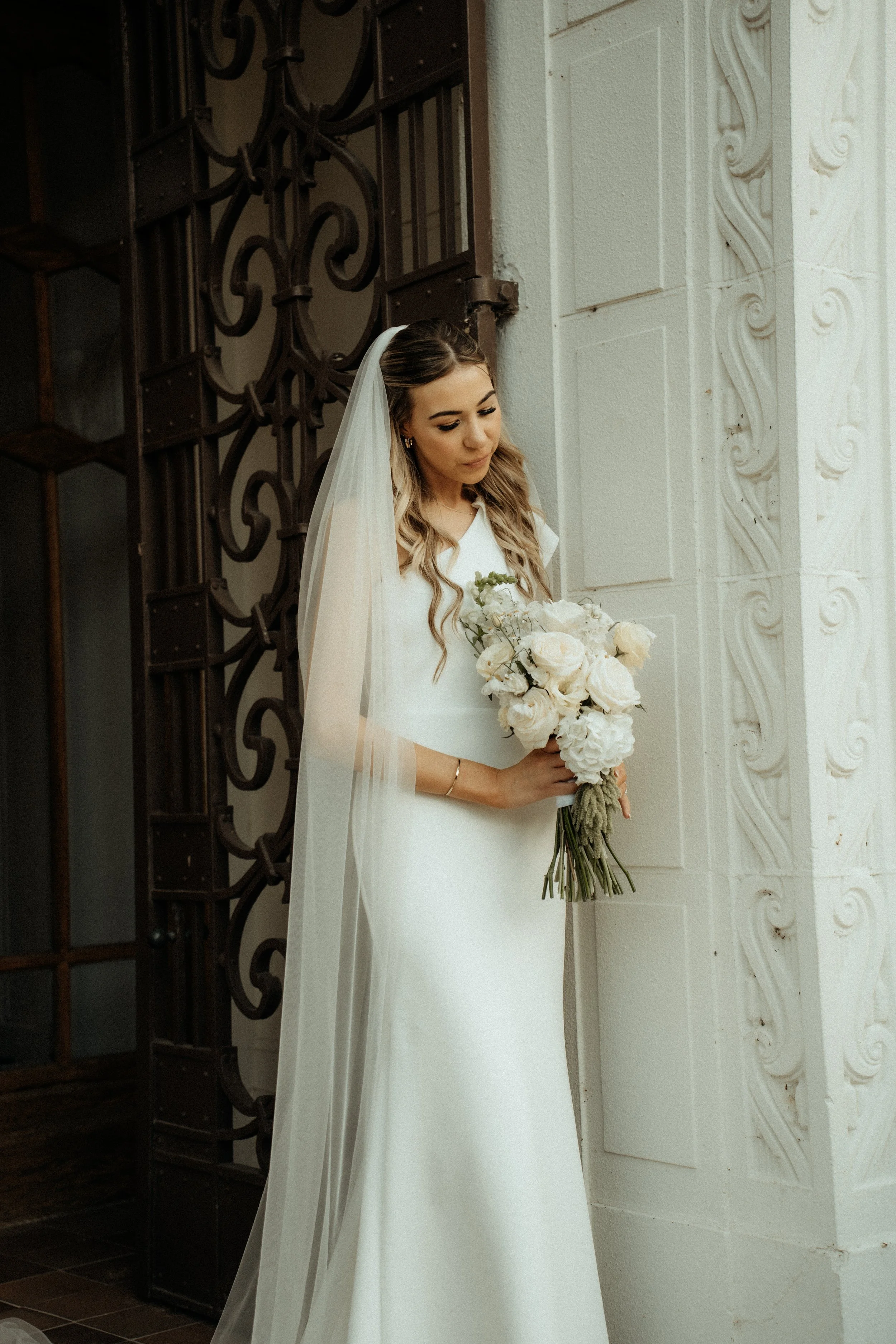 A bride in a white wedding dress and veil holding a bouquet of white roses, standing indoors near a white decorative wall with swirling patterns.