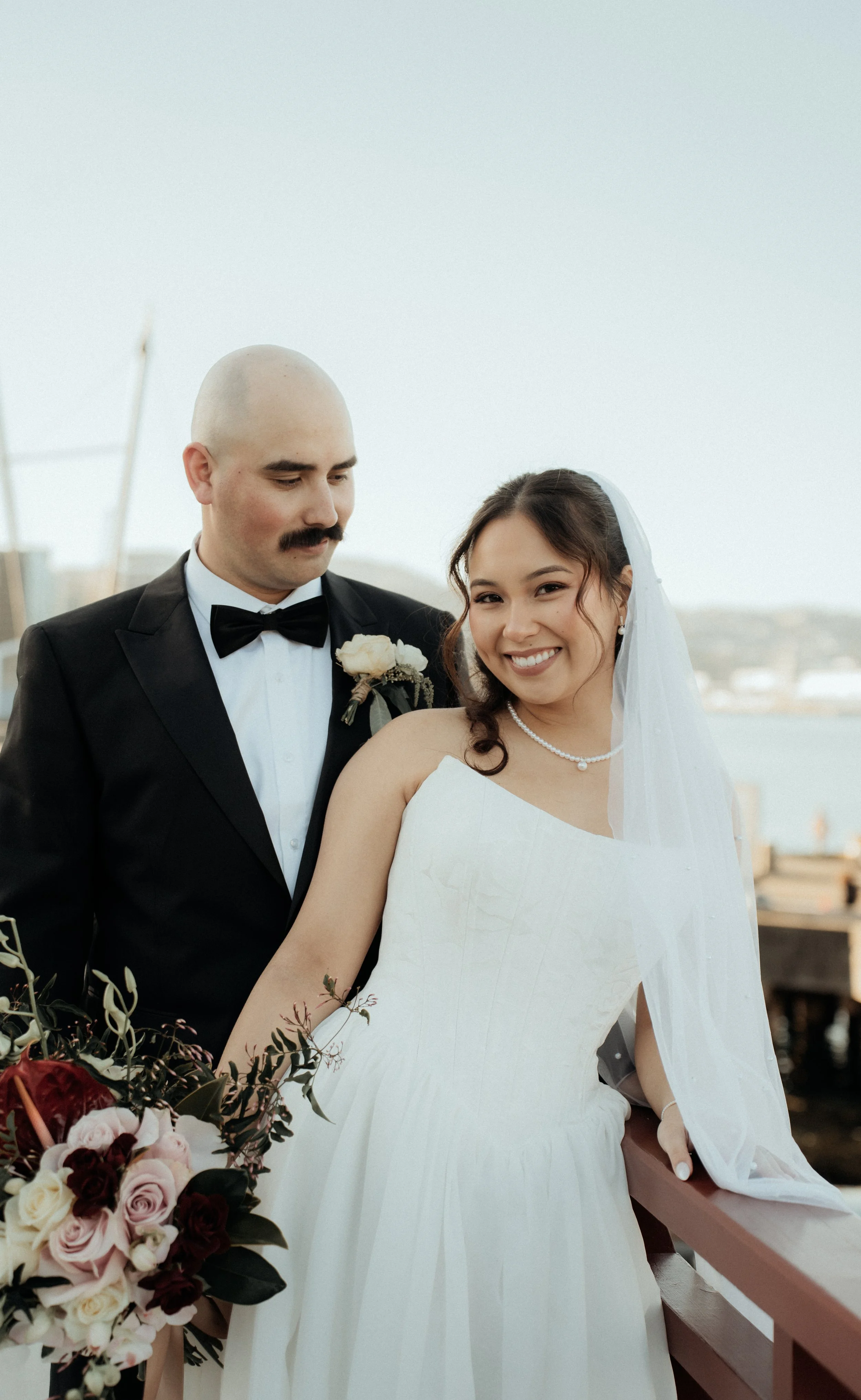 A bride and groom on their wedding day, standing outdoors on a bridge or balcony. The bride is smiling, wearing a white wedding dress and veil, with a pearl necklace. The groom, with a bald head and mustache, is dressed in a black tuxedo with a bow t