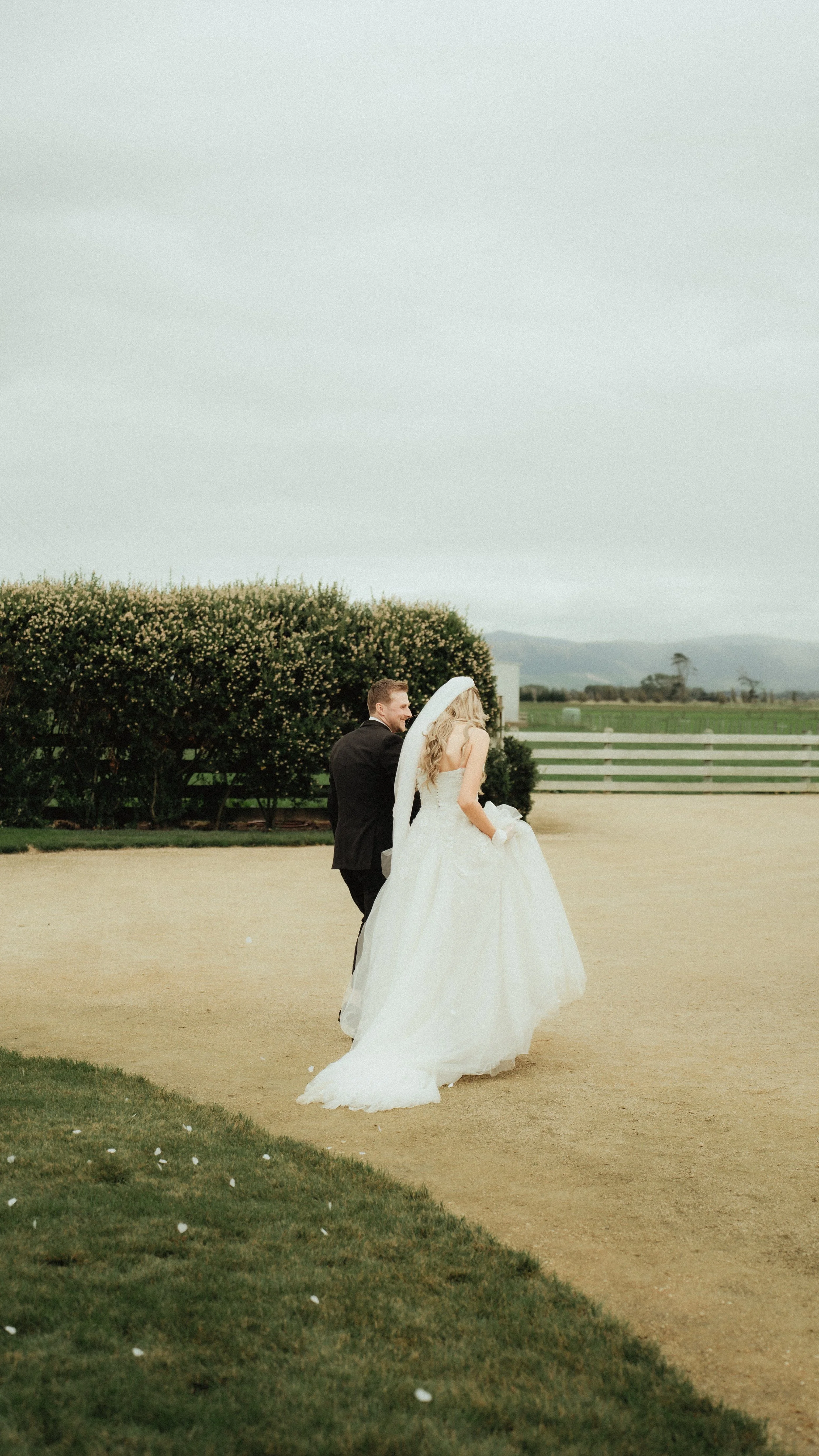 A bride and groom walking together outdoors on a dirt path, with the bride in a white wedding gown and veil, and the groom in a black suit, in a scenic rural setting with trees, a white fence, and mountains in the background.