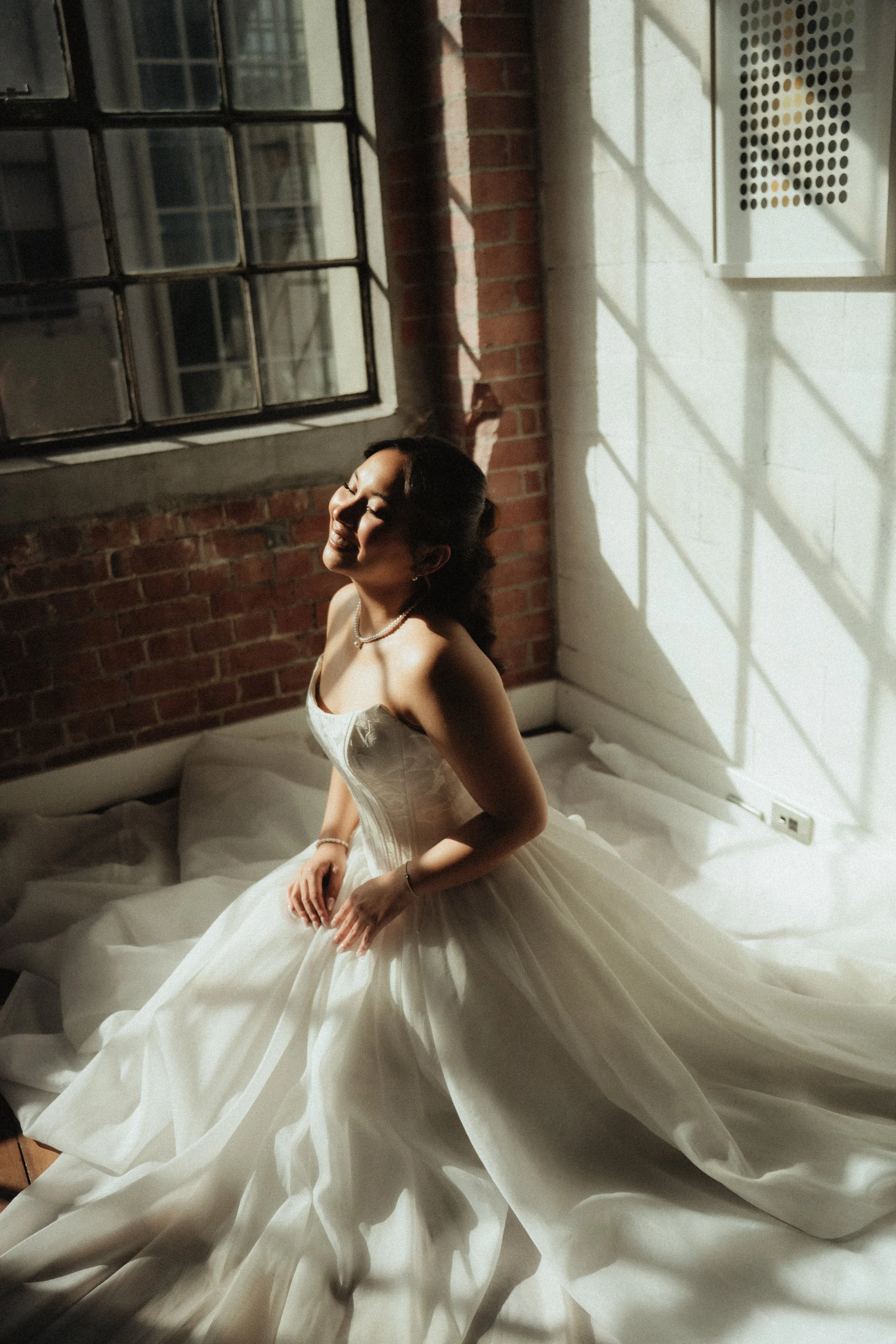 A woman in a white wedding dress sitting on a bed, smiling with her eyes closed, sunlight casting shadow patterns on the wall behind her.