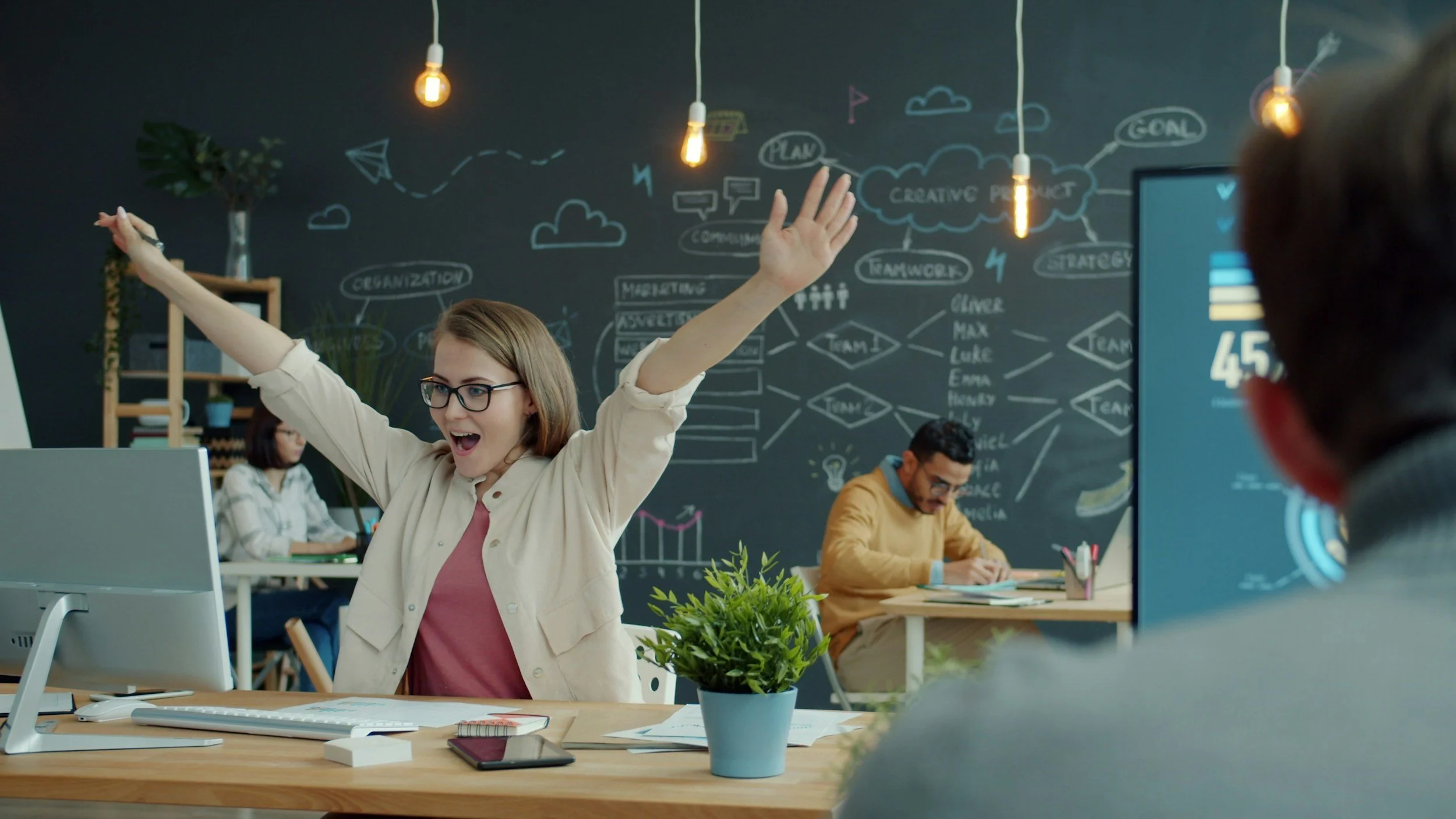A woman in glasses and a beige shirt raising her arms and celebrating in a modern office with colleagues working at desks and a chalkboard with diagrams and words like 'Organization,' 'Marketing,' and 'Creative Product' in the background.