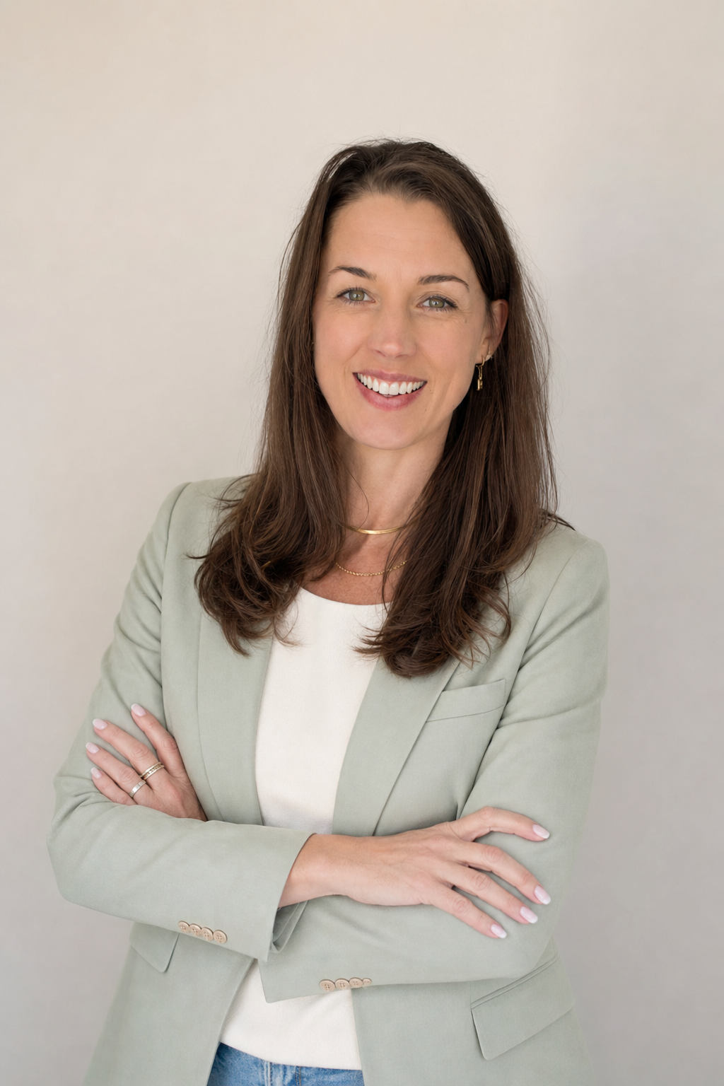 Joelene Edwards with brown hair and green eyes, smiling, wearing a light green blazer over a white top, standing with arms crossed in front of a plain white background.