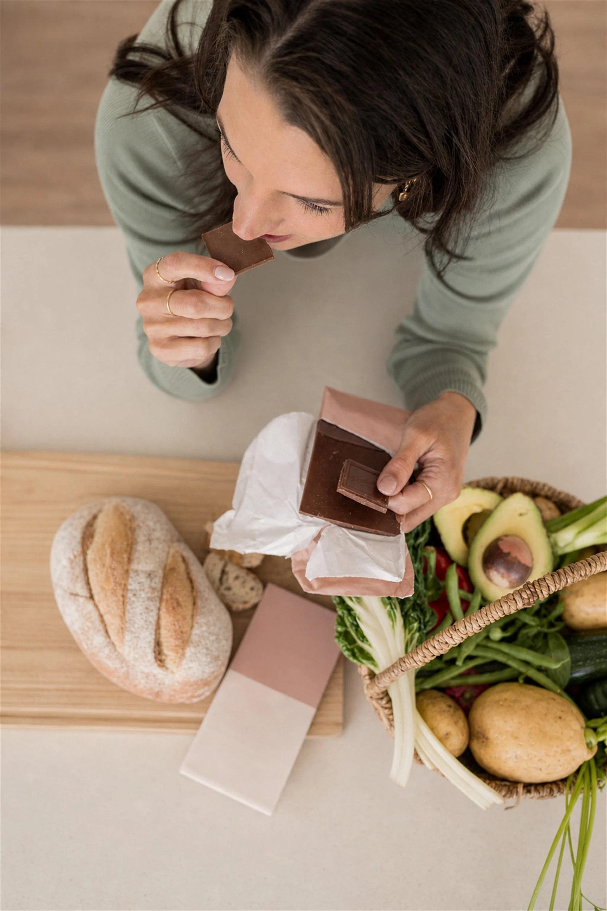 Top-down view of a woman with dark hair eating a piece of chocolate, with bread, a grocery bag with vegetables and potatoes, and sliced avocado on a wooden table.