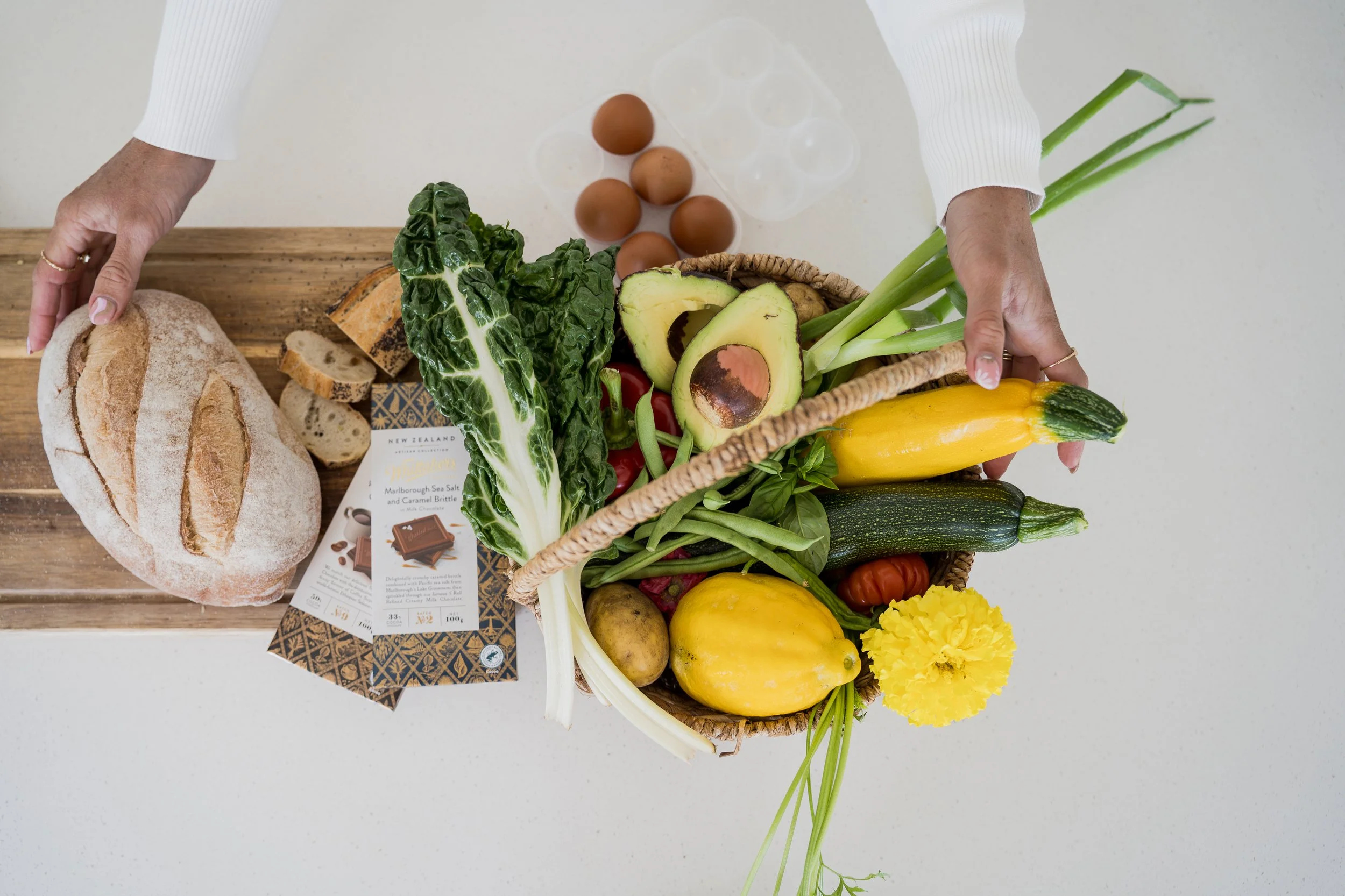 A top-down view of a white surface with a wicker basket filled with various fresh vegetables, including avocados, yellow zucchini, green onions, and cherry tomatoes. To the left on a wooden surface, there is a loaf of rustic bread and a chocolate bar package.
