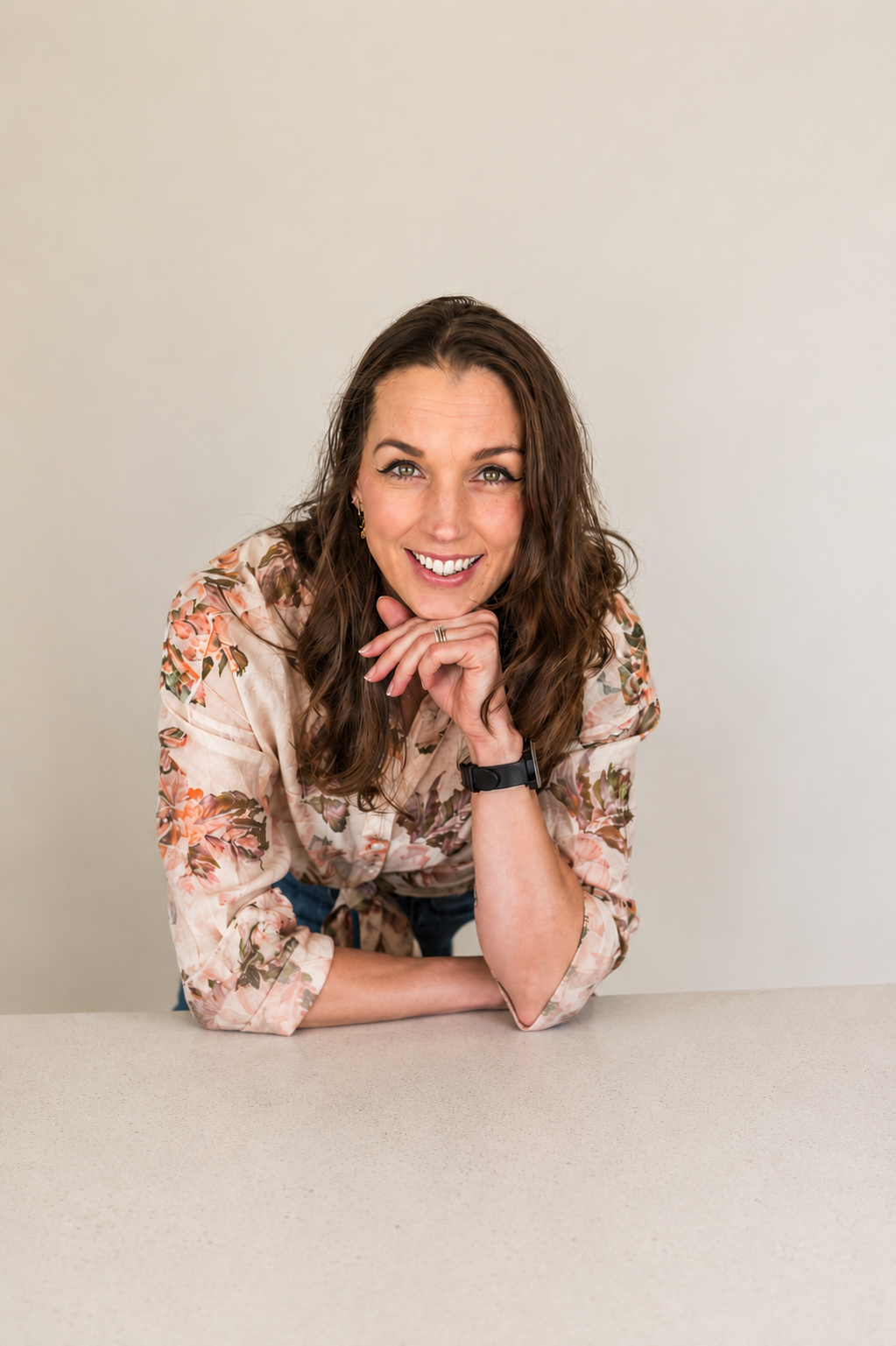 Joelene Edwards with long, wavy brown hair sitting at a beige table against a plain off-white background. She is smiling, resting her chin on her hand, and wearing a floral blouse with rolled-up sleeves, a black watch, and earrings.