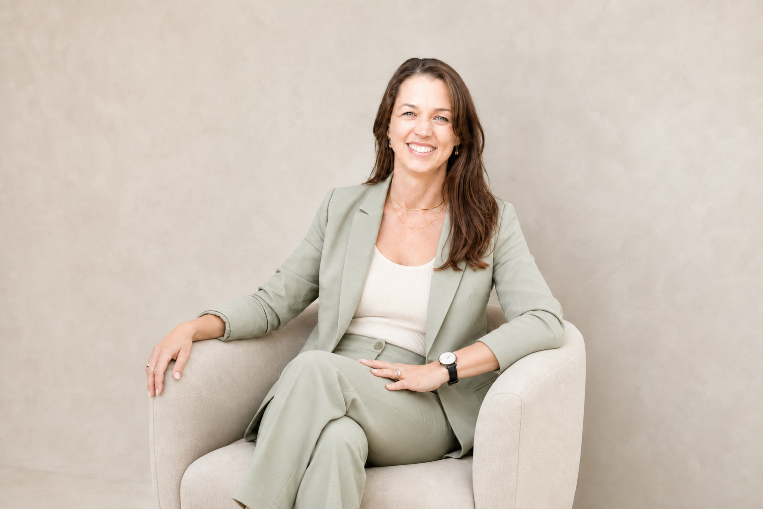 Joelene Edwards with long brown hair smiling, sitting on a beige armchair against a neutral background, wearing a light green suit with a white top, gold jewelry, and a watch.