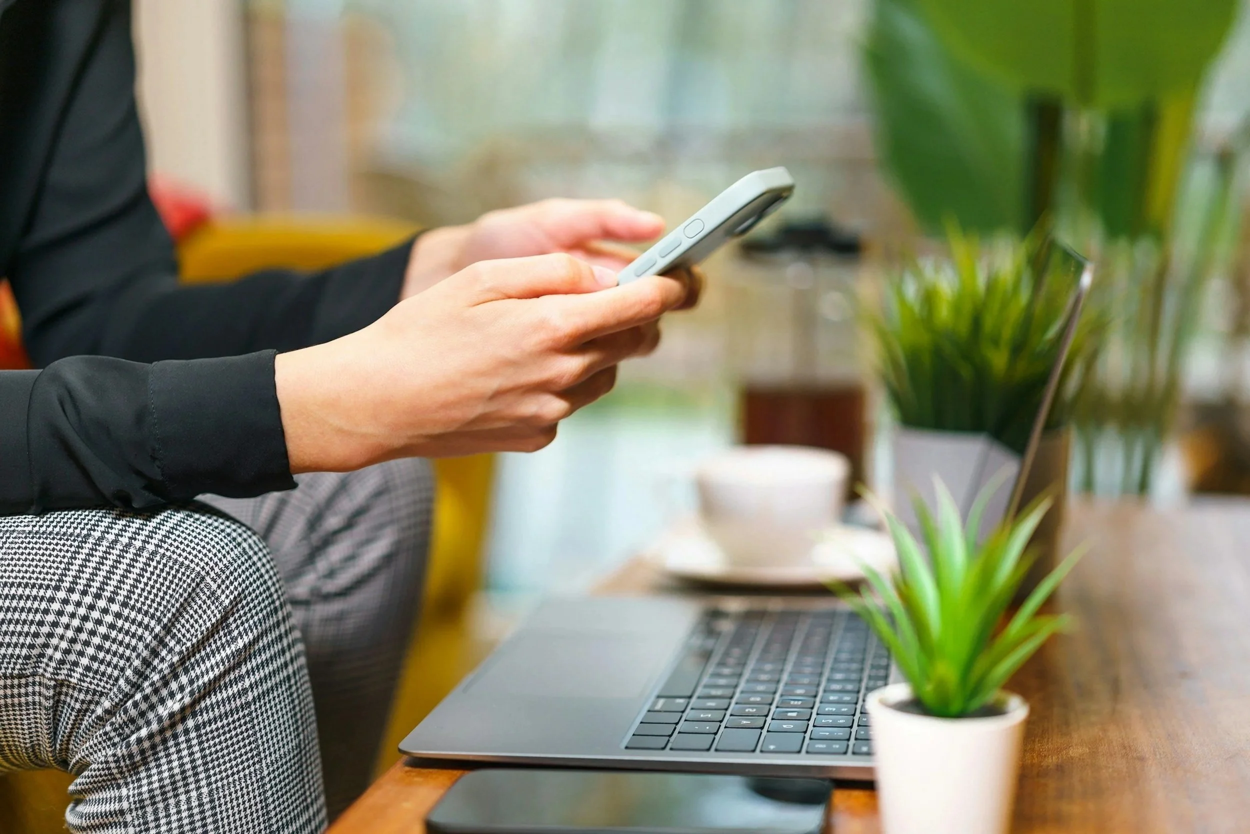 Person using a smartphone at a desk with a laptop, plant, and coffee mug nearby in a bright, indoor setting.
