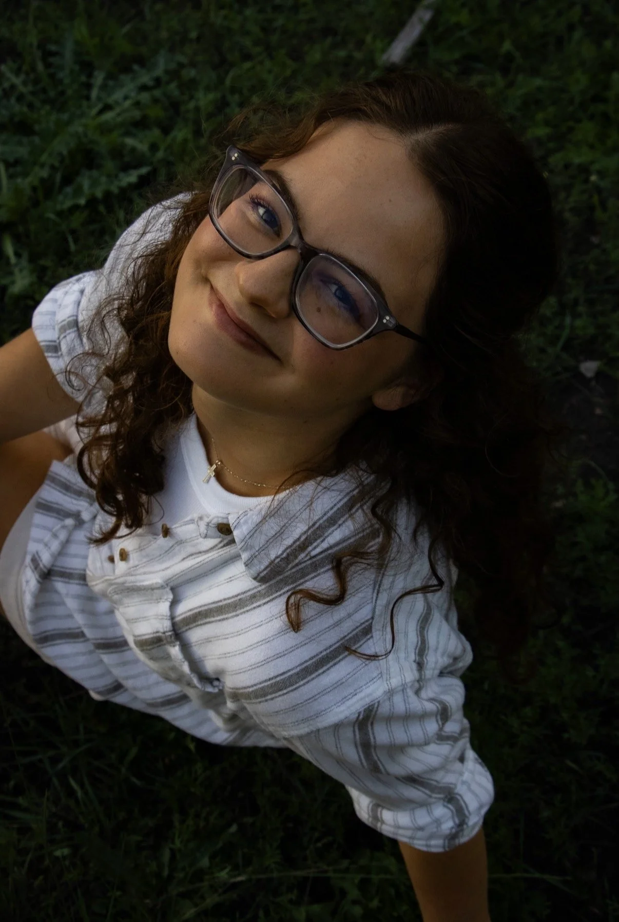 A woman with curly brown hair, glasses, and freckles lies on her back on green grass, smiling at the camera, wearing a striped button-up shirt and a delicate necklace.