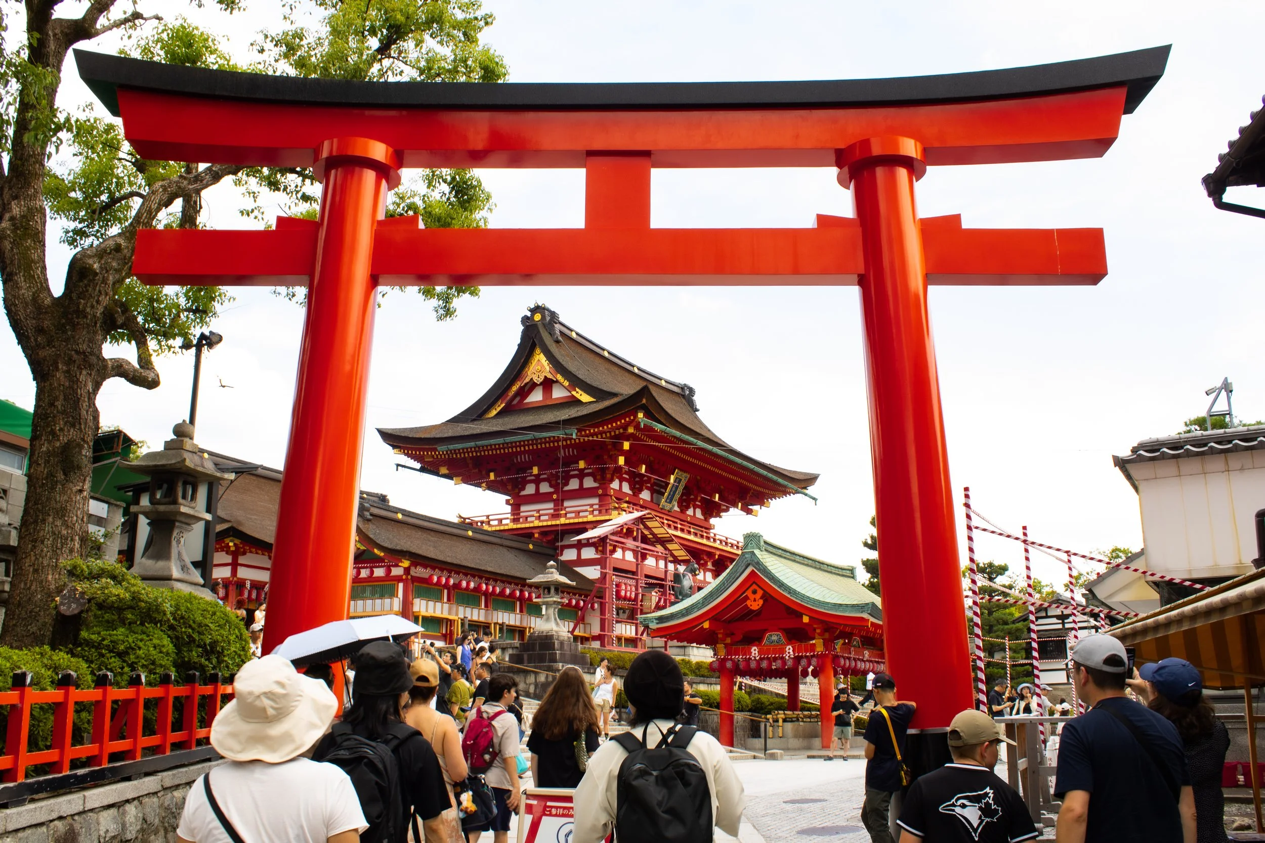 Traditional Japanese shrine entrance with a large red torii gate and a temple behind it, surrounded by people visiting and walking through the area.
