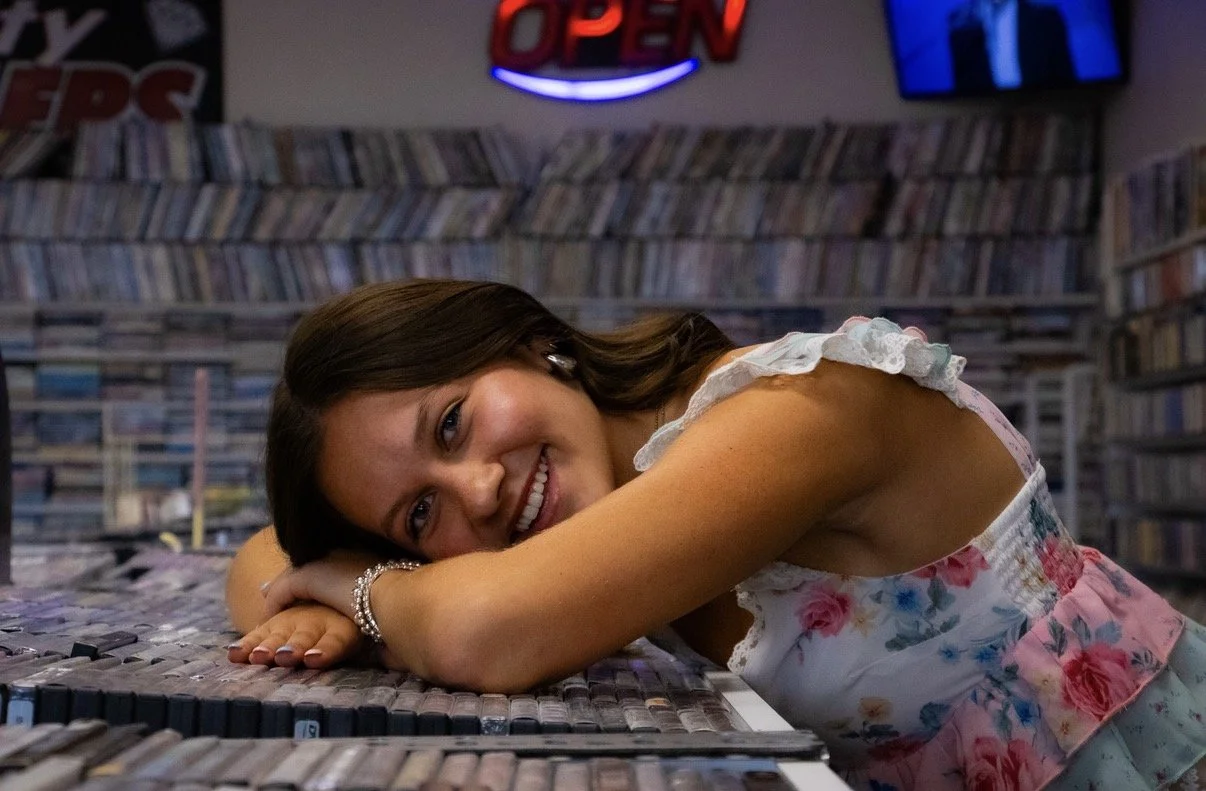 A young woman with brown hair and a floral dress is smiling and resting her head on her folded arms on a table in a bookstore or library, with shelves of books behind her and an illuminated 'Open' sign above.