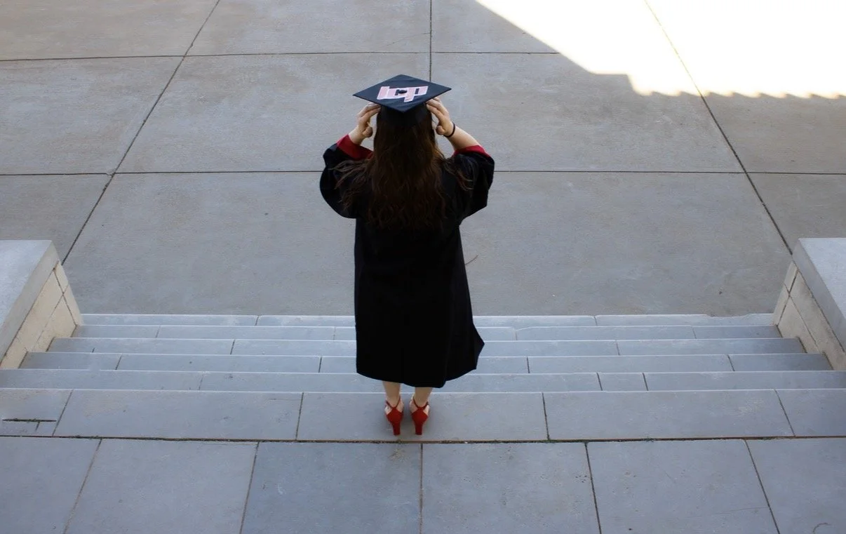 A woman in a graduation cap and gown standing on the steps of a building, viewed from above.