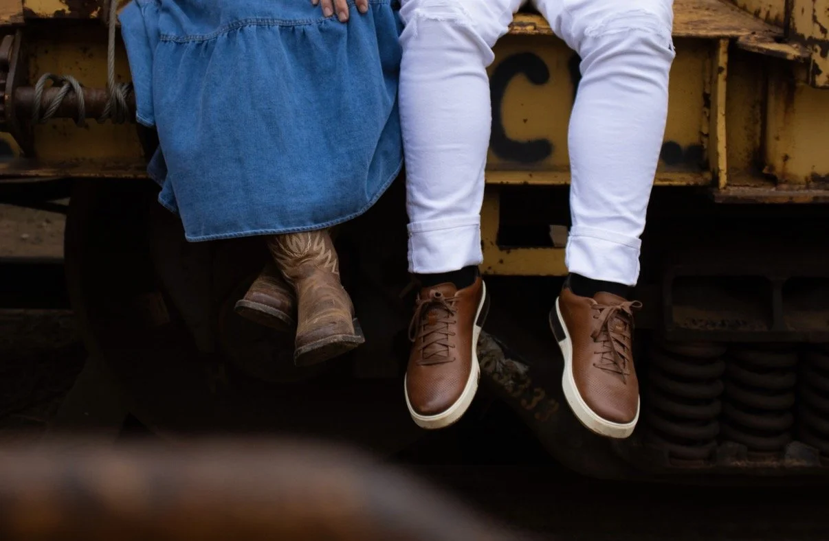 Two children sitting on a yellow train, with only their legs and shoes visible. One child is wearing brown sneakers and white pants, and the other is wearing brown cowboy boots and a blue dress.