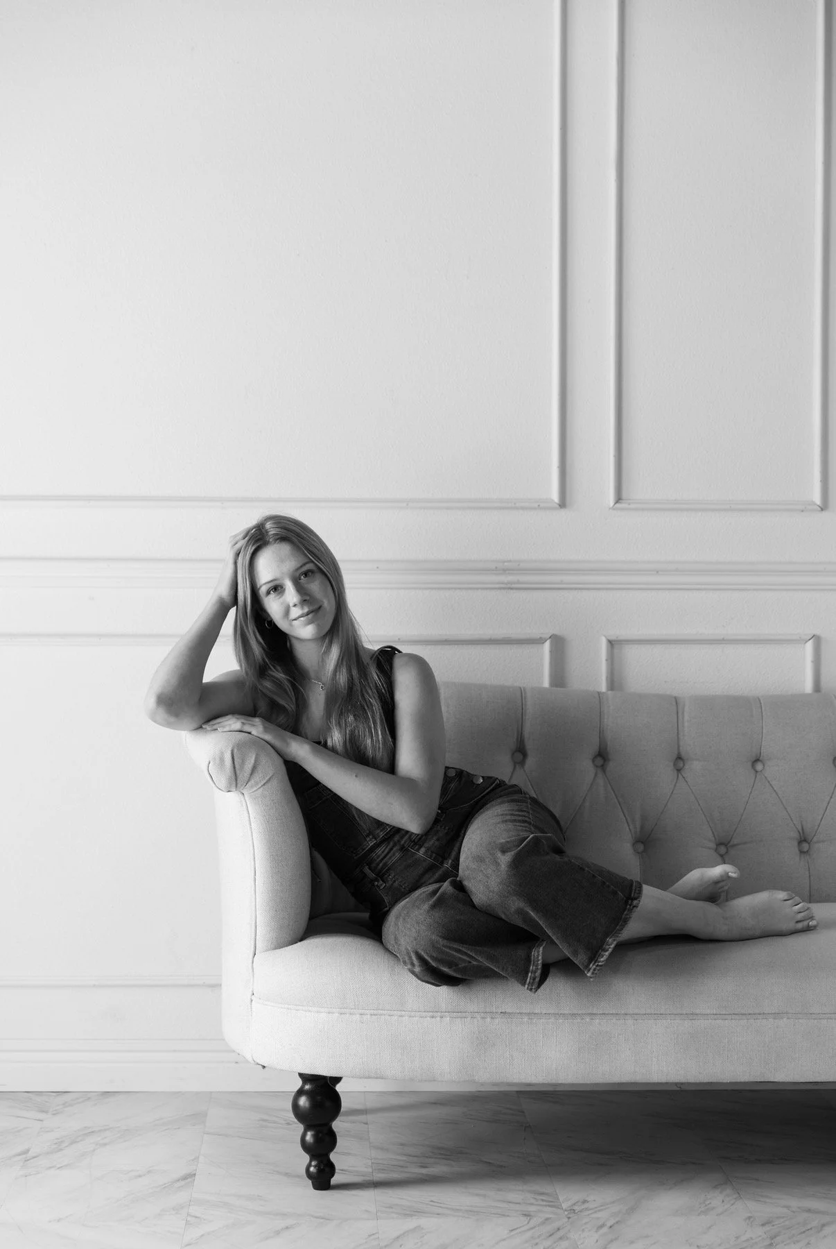 A young woman with long hair is lying on a vintage-style sofa with tufted upholstery, one arm resting on the back and her hand touching her hair, in a room with decorative wall paneling. The photo is in black and white.