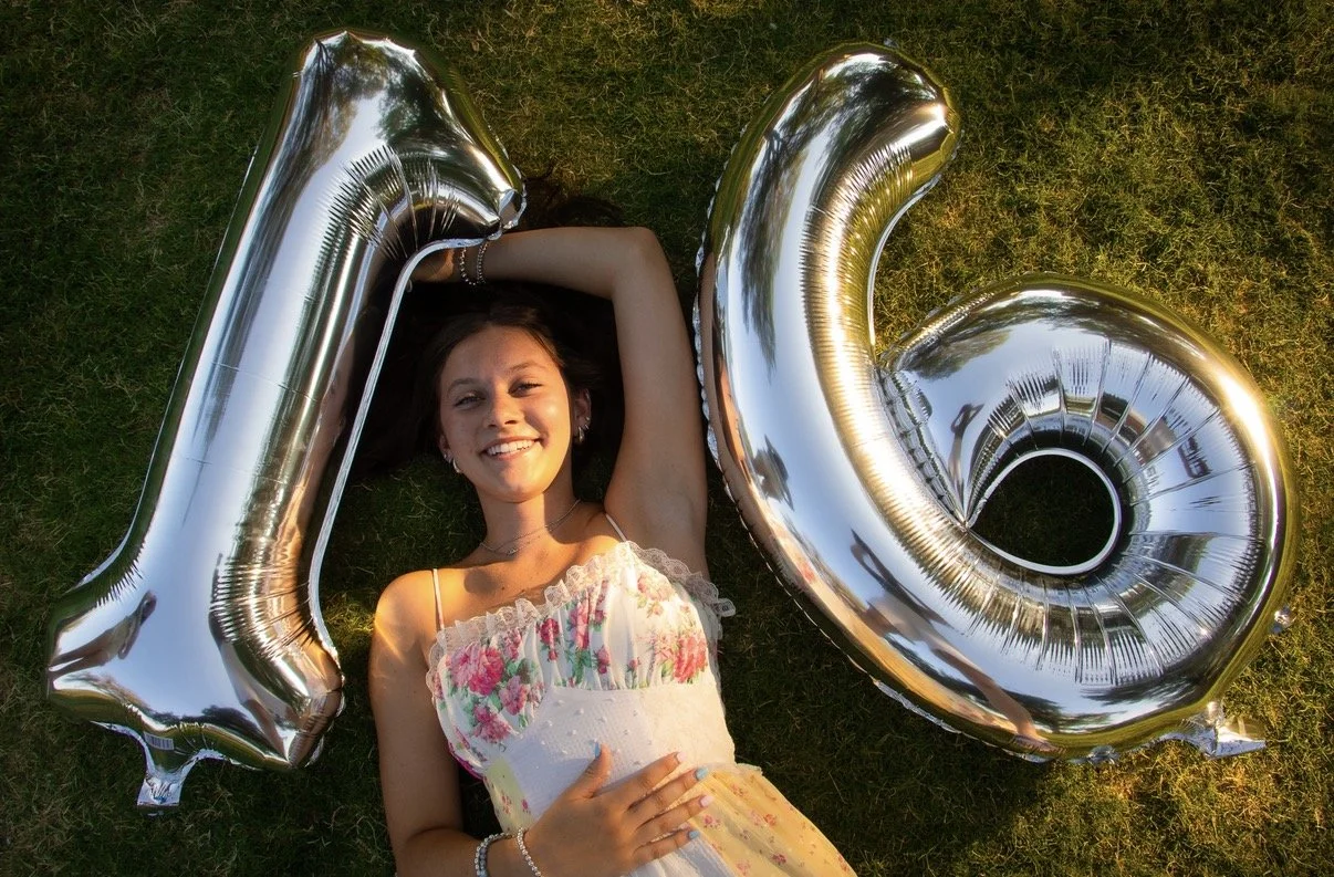 A young woman lying on grass with a big smile, holding a large metallic number 1 and a large metallic number 0, celebrating a special occasion.