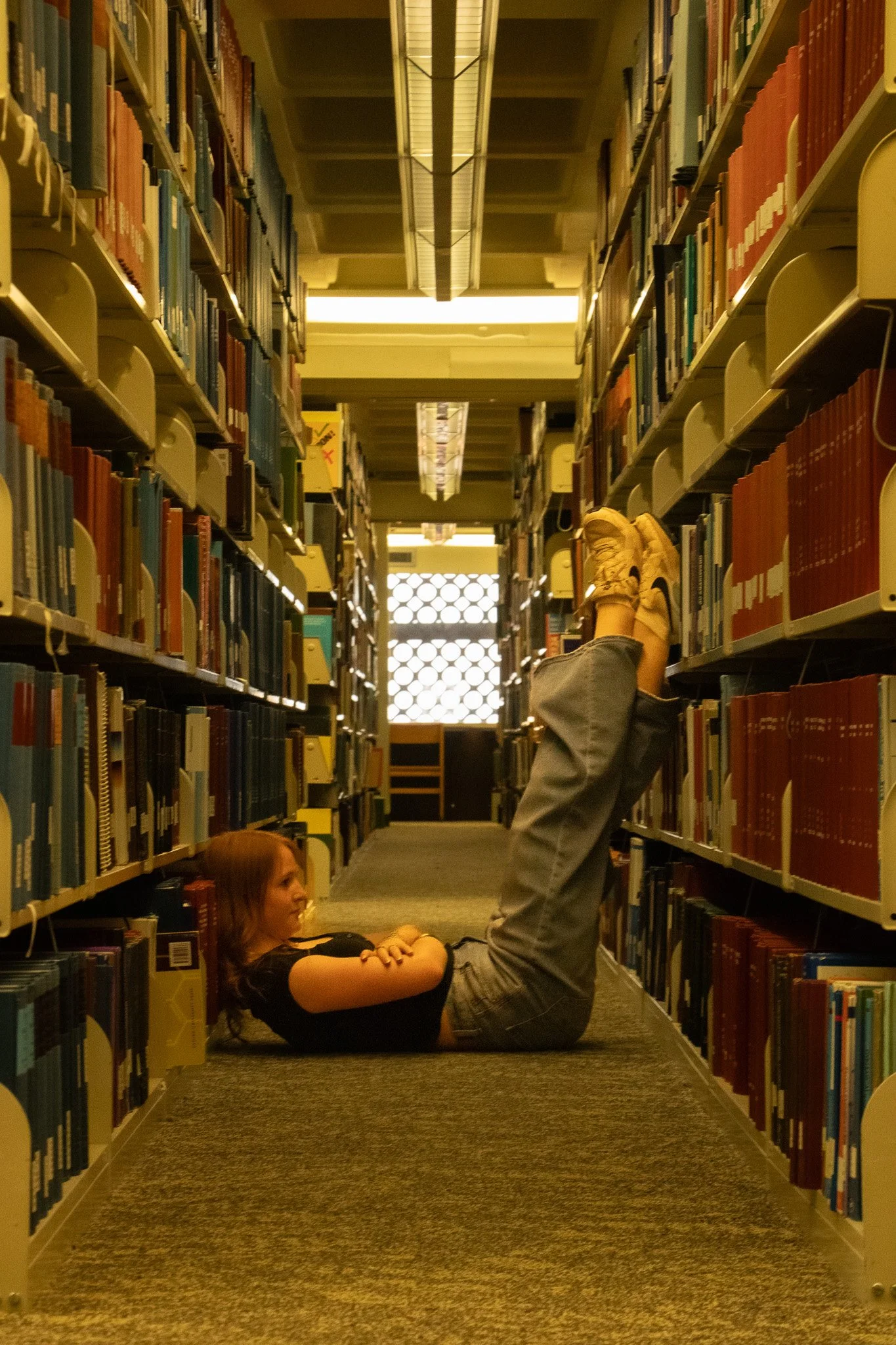 A girl lying on her back on a library floor between bookshelves, with her head resting on the floor and legs propped up on the bookshelf, doing a playful pose. A boy is standing nearby with his feet resting on the bookshelves, also in a playful pose. The scene is inside a library.