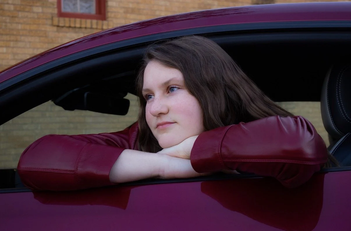 A young woman with brown hair and blue eyes, wearing a burgundy leather jacket, is sitting in a car with her arms resting on the window ledge.