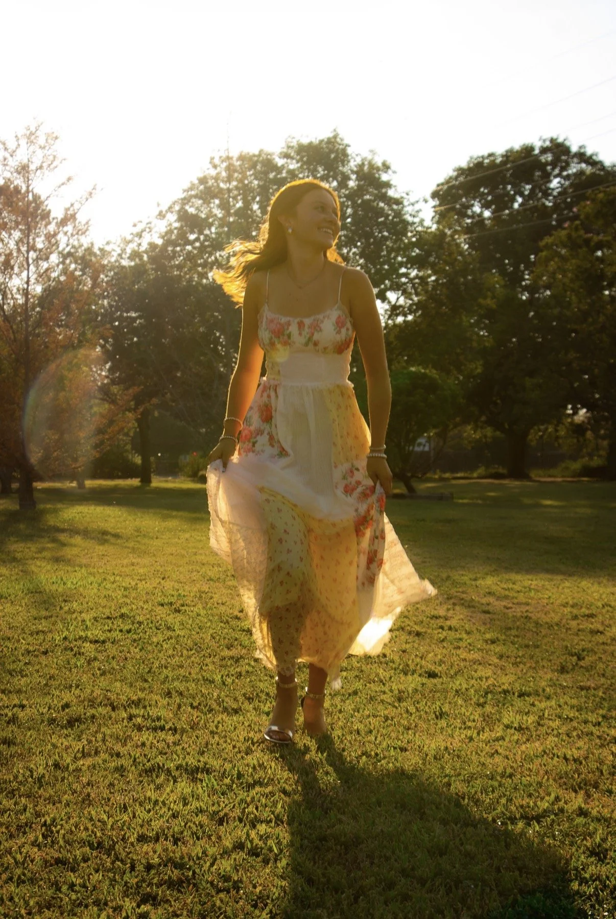 A woman in a floral dress walking on a grassy field during sunset with trees in the background.