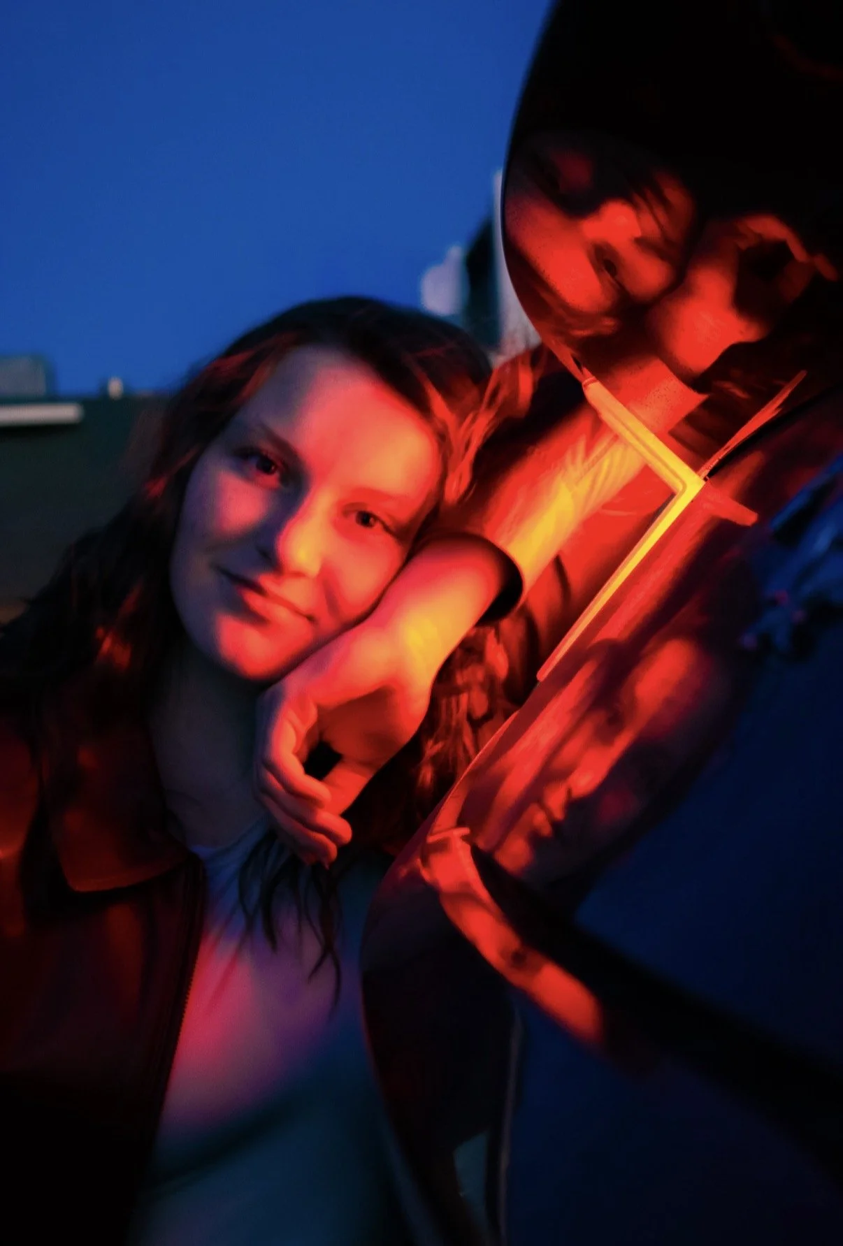 A young woman with long brown hair, smiling, resting her head on her hand, next to a person wearing glasses and a jacket with glowing neon accents, in an outdoor setting during dusk or night.