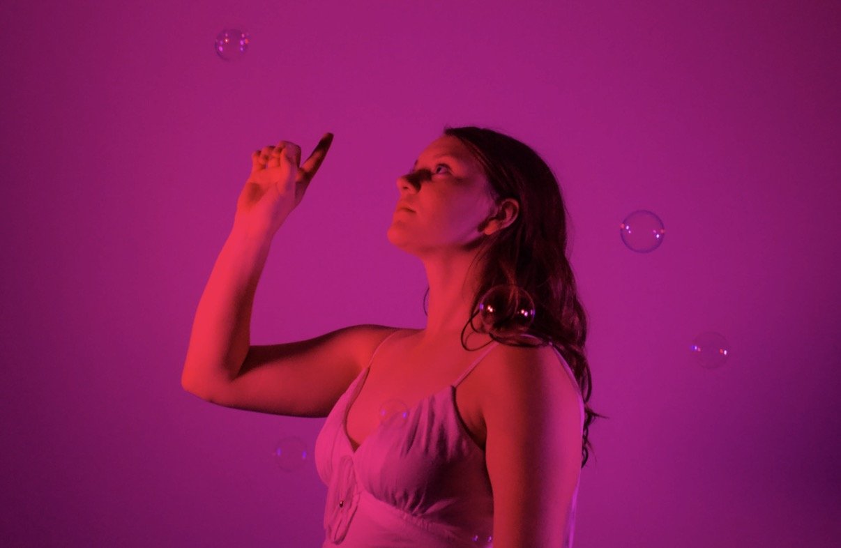 A woman with long hair wearing a sleeveless top, surrounded by bubbles, illuminated with pink and purple lighting.