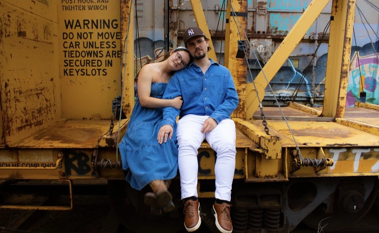 A young couple sitting on a yellow industrial train car with graffiti in the background. The woman is leaning on the man's shoulder, both dressed in blue and white clothing, with the woman in a blue dress and the man in a blue shirt and white pants.