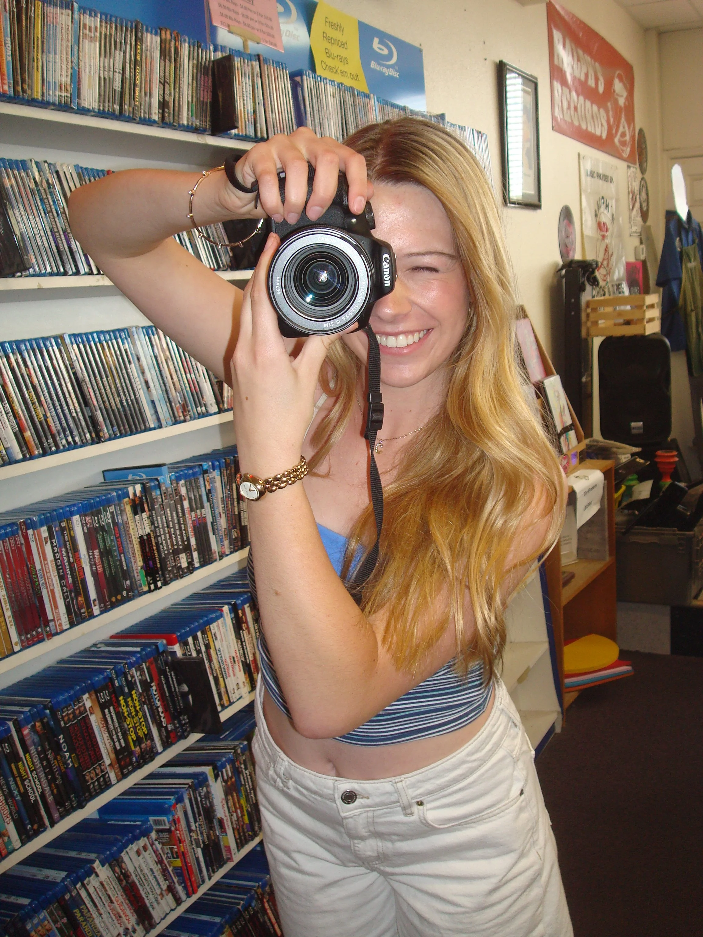 A woman with long red hair taking a photo using a Canon camera in a store with shelves filled with DVDs and Blu-ray discs. She is smiling, wearing a striped blue and white sleeveless top and white shorts, with jewelry on her wrist and neck.