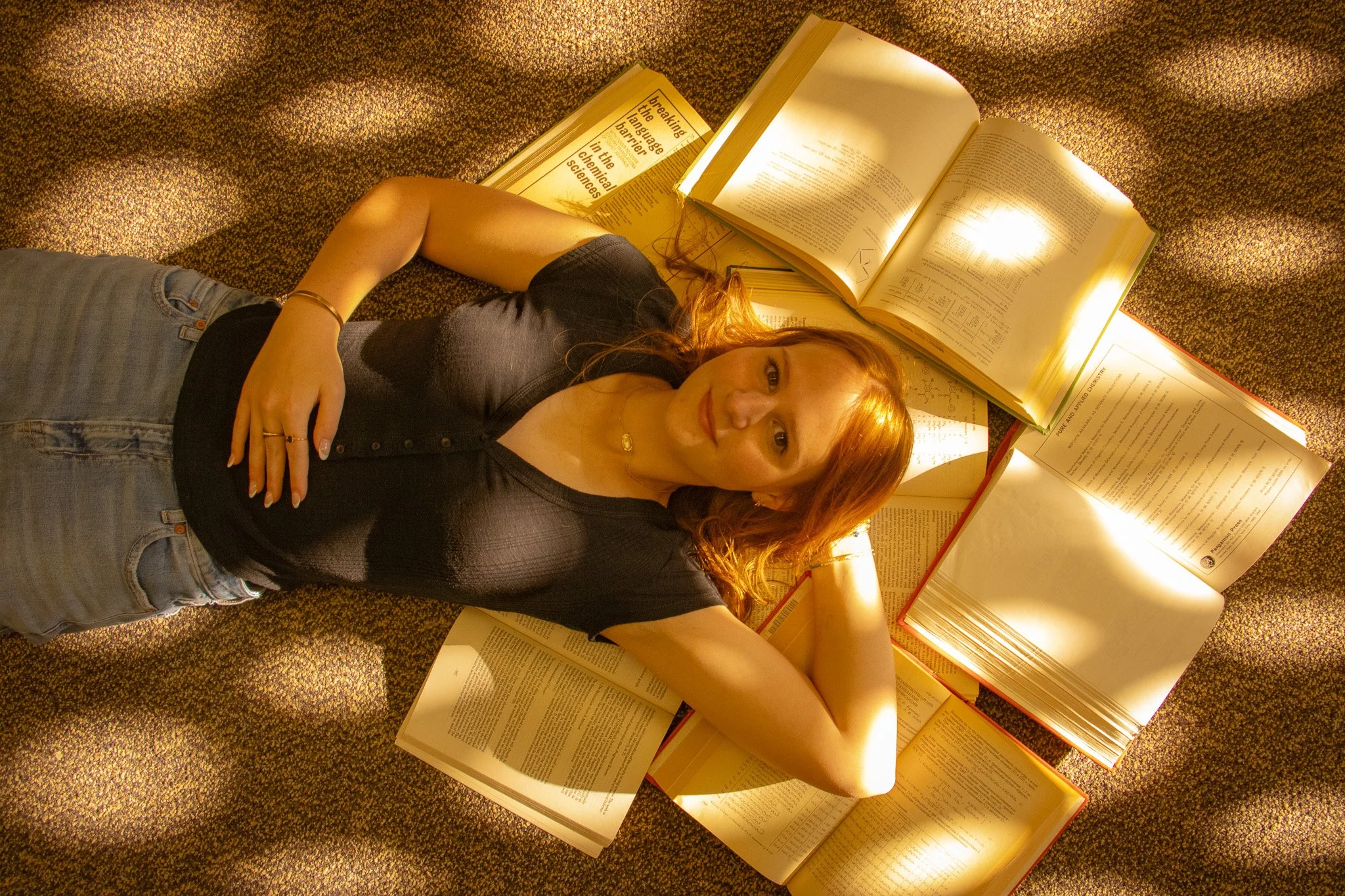 A woman with red hair lying on a patterned carpet surrounded by open textbooks and study materials, looking up at the camera.
