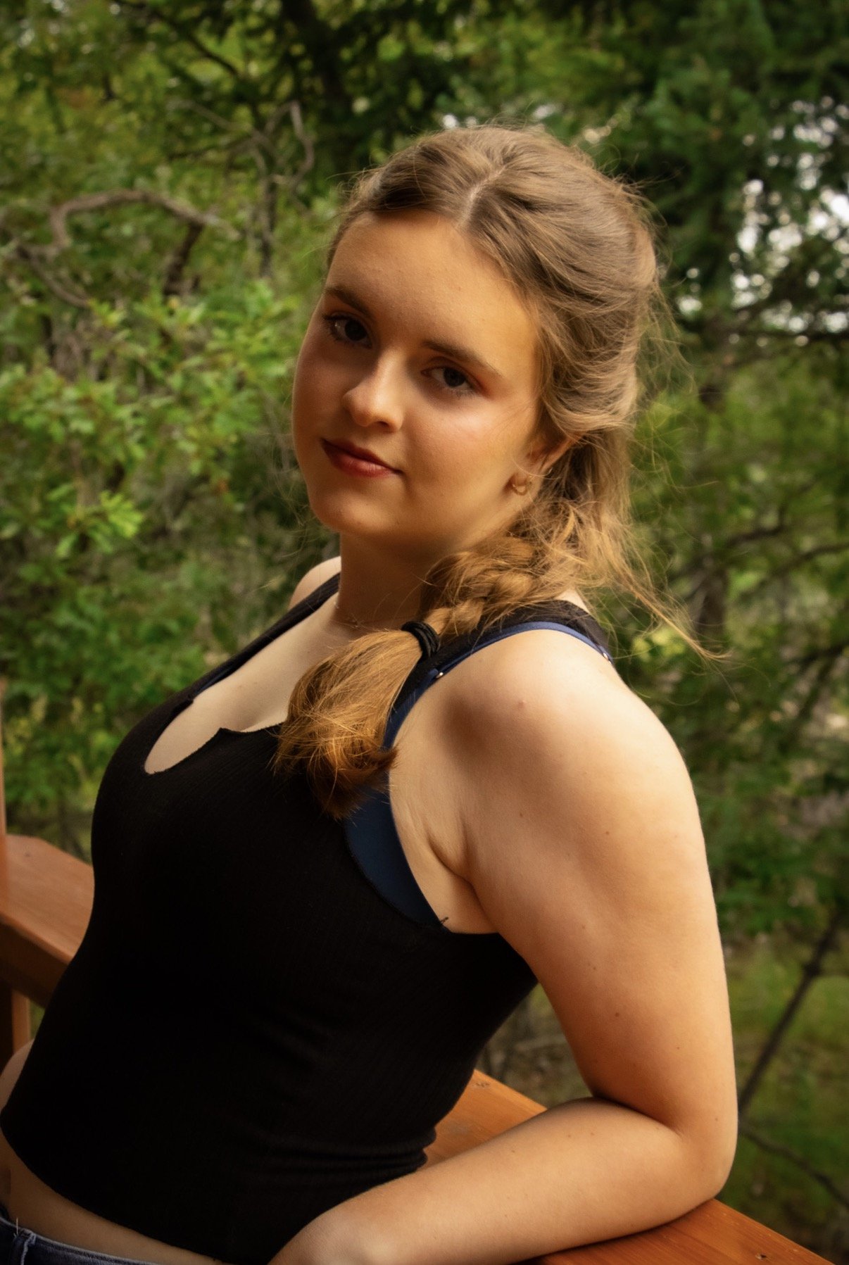 A young woman with light brown hair in a braid, wearing a black tank top with blue straps, standing outdoors on a wooden deck with green trees in the background.