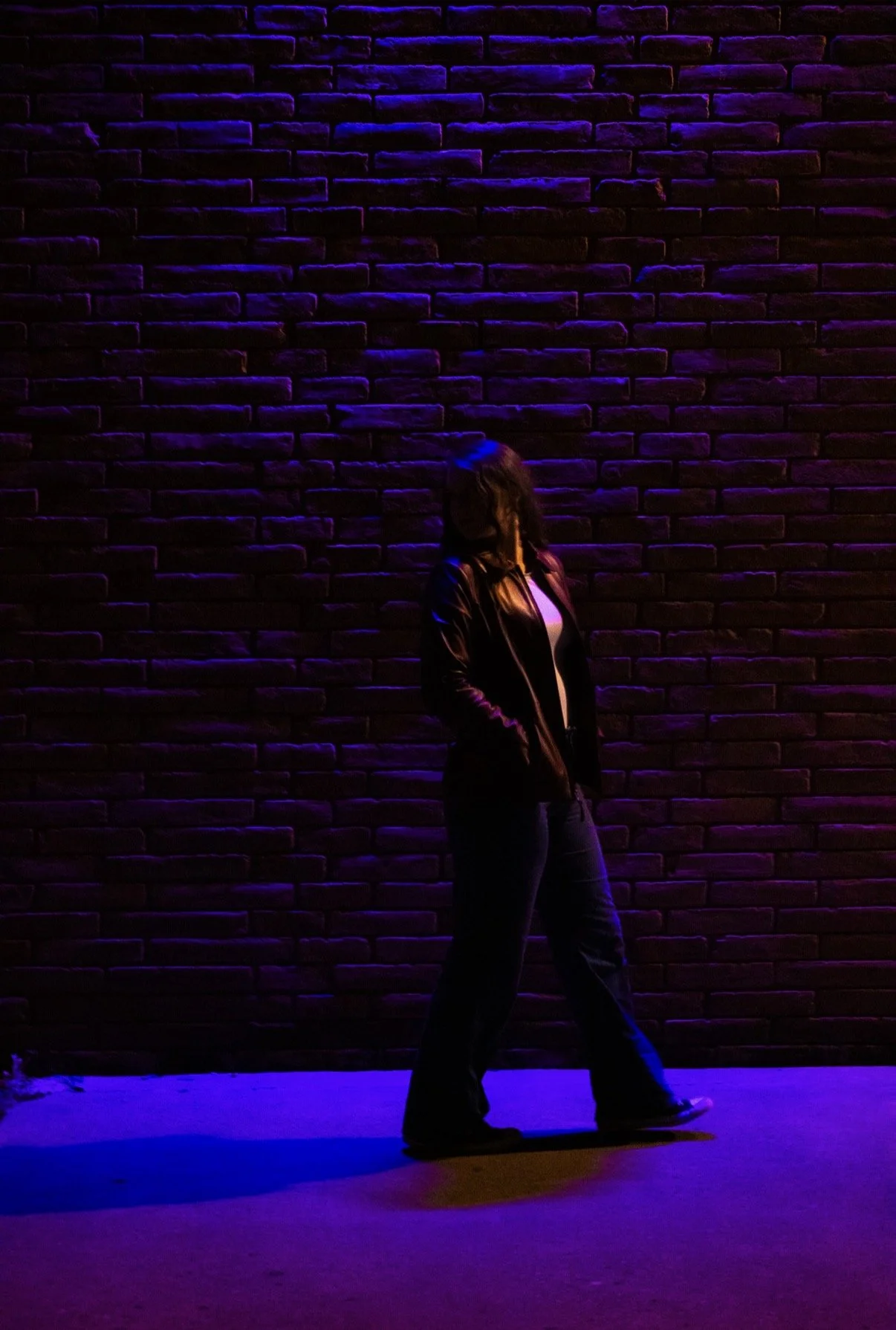 A person stands against a dark brick wall illuminated with purple and blue neon lights, wearing a jacket and jeans, with hands in pockets and head tilted downward.