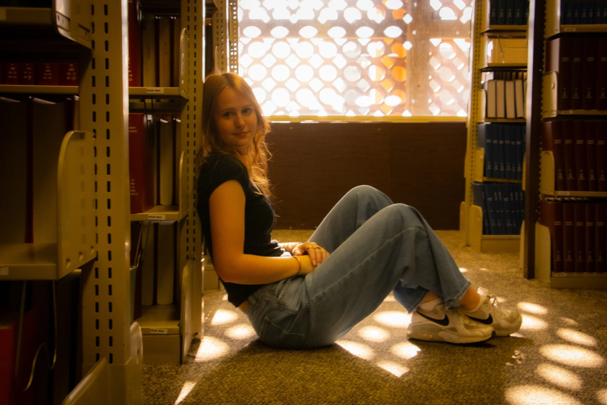 A young woman with blonde hair sitting on the floor of a library between bookshelves, backlit by sunlight coming through a patterned window, wearing a black t-shirt, jeans, and sneakers.