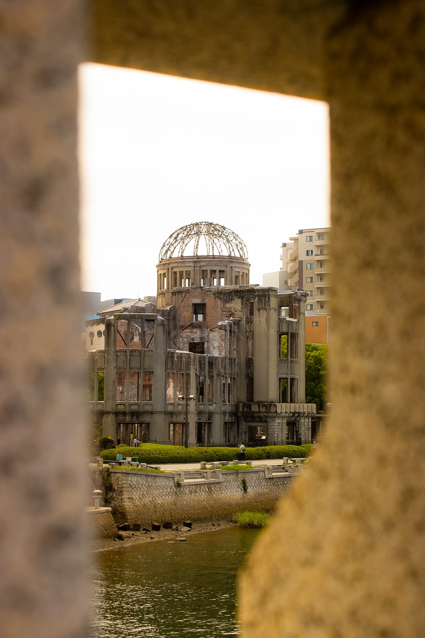 View of the A-Bomb Dome, Hiroshima, Japan, seen through a small opening between a brick wall and a concrete wall, with the river below and people walking along the park.